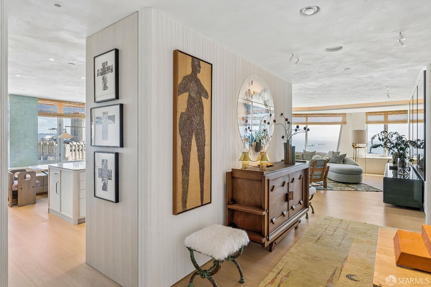This sophisticated interior view captures a transition space featuring a textured wall adorned with a tall, abstract figure painting and a gallery of three framed cross-motif artworks. To the right, a wooden sideboard sits beneath a circular mirror, while a plush, fur-topped stool adds a touch of texture to the foreground. The perspective looks through the open-plan layout, revealing a bright kitchen on the left and a sun-drenched living area with large windows overlooking the ocean in the distance.