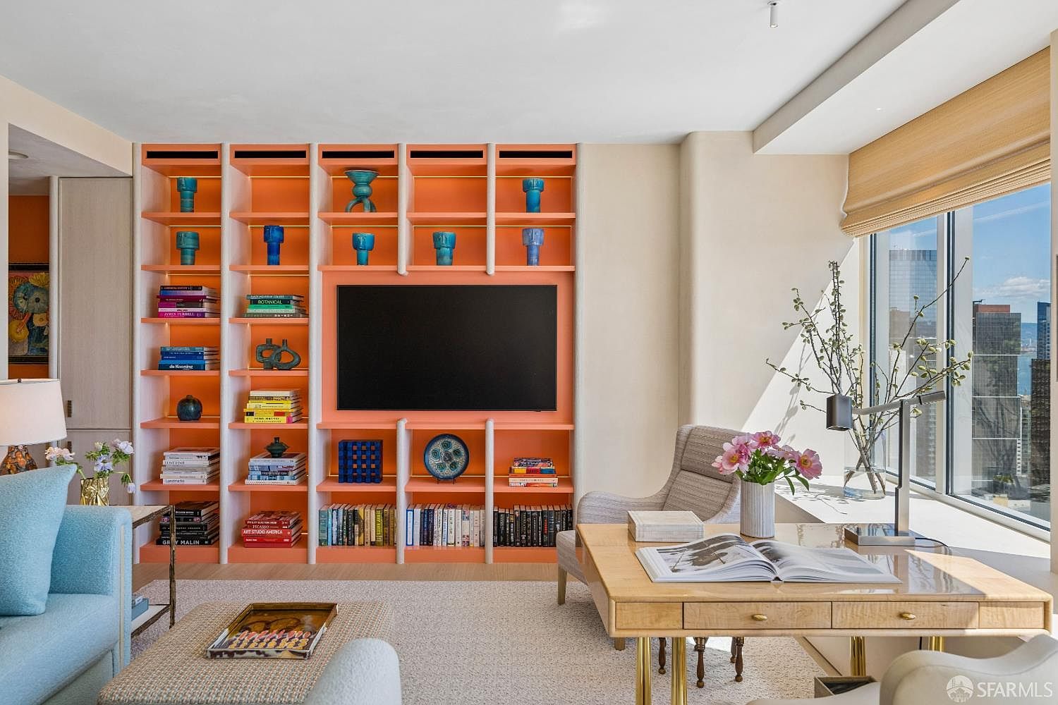 This bright and contemporary living room features a striking, custom-built coral-colored shelving unit that serves as a focal point, housing a television, books, and decorative blue vases. To the right, a light wood desk sits near large windows that offer an expansive city view, while a plush blue sofa is positioned in the foreground. The space is filled with natural light, creating an inviting and sophisticated atmosphere suitable for both relaxation and work.