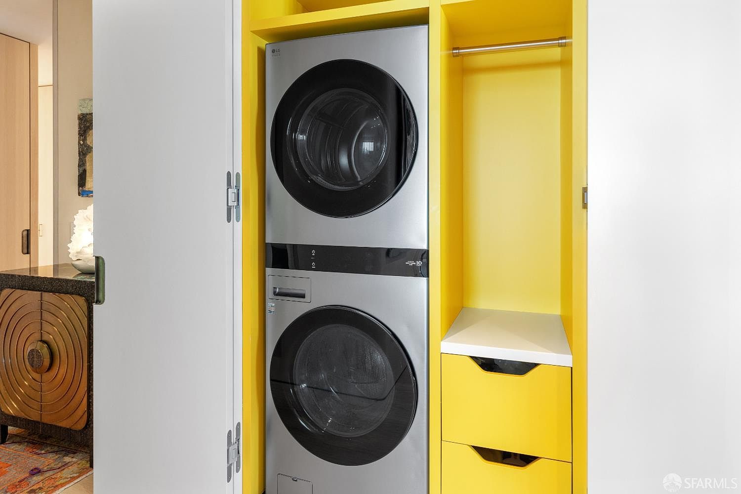 This laundry area features a stacked LG washer and dryer unit set within a vibrant, bright yellow built-in cabinet structure. To the right of the appliances, there is an open closet space with a hanging rod and two drawers below, providing organized storage. The clean, modern design is accented by white cabinetry and a white countertop, creating a functional and visually striking utility space.