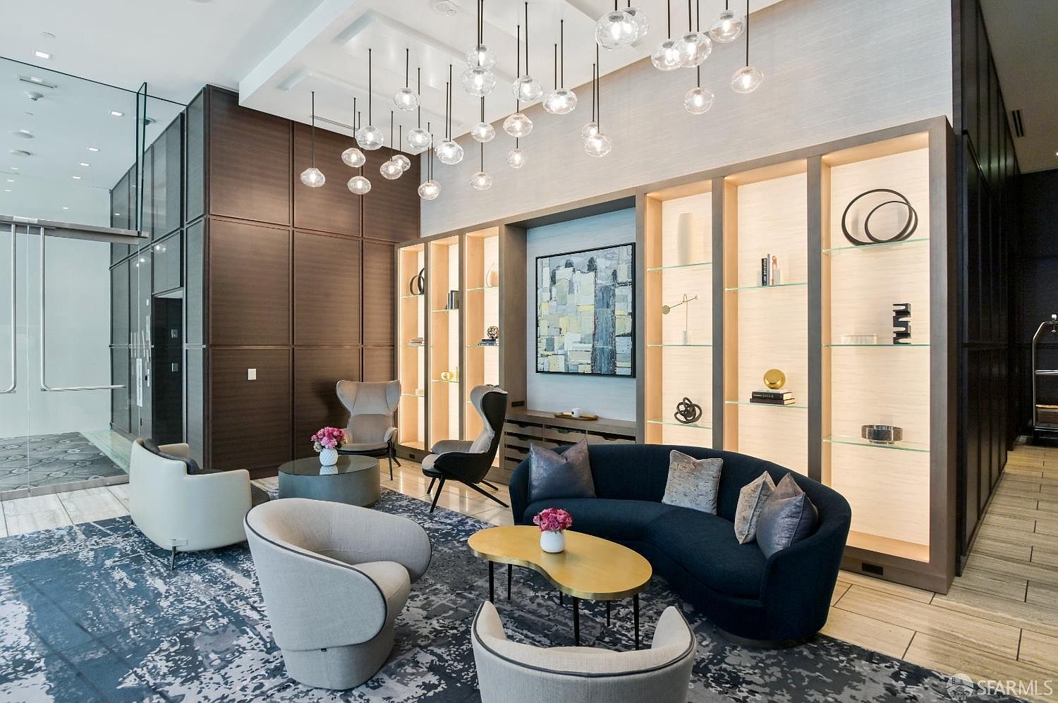 This sophisticated lobby or lounge area features a contemporary design with a dark, curved sofa and several plush armchairs arranged on a patterned area rug. The space is anchored by a large, illuminated shelving unit with glass display cases and a striking abstract painting, all beneath a dramatic installation of numerous spherical pendant lights. The perspective is a wide, eye-level shot that captures the elegant, high-end atmosphere of a modern residential building's common area.