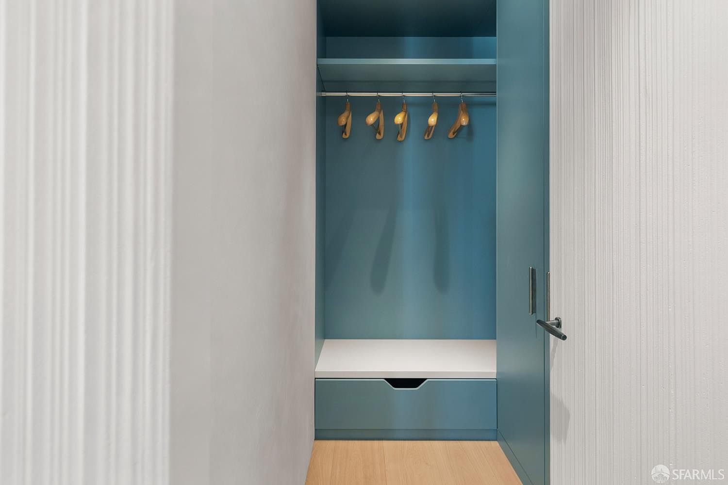 This modern walk-in closet features a striking teal-colored built-in storage unit with a hanging rod, a shelf, and a lower drawer. The space is accented by a white bench seat and light wood flooring, creating a clean and organized aesthetic. The perspective is a straight-on view looking into the closet, highlighting its compact and functional design.