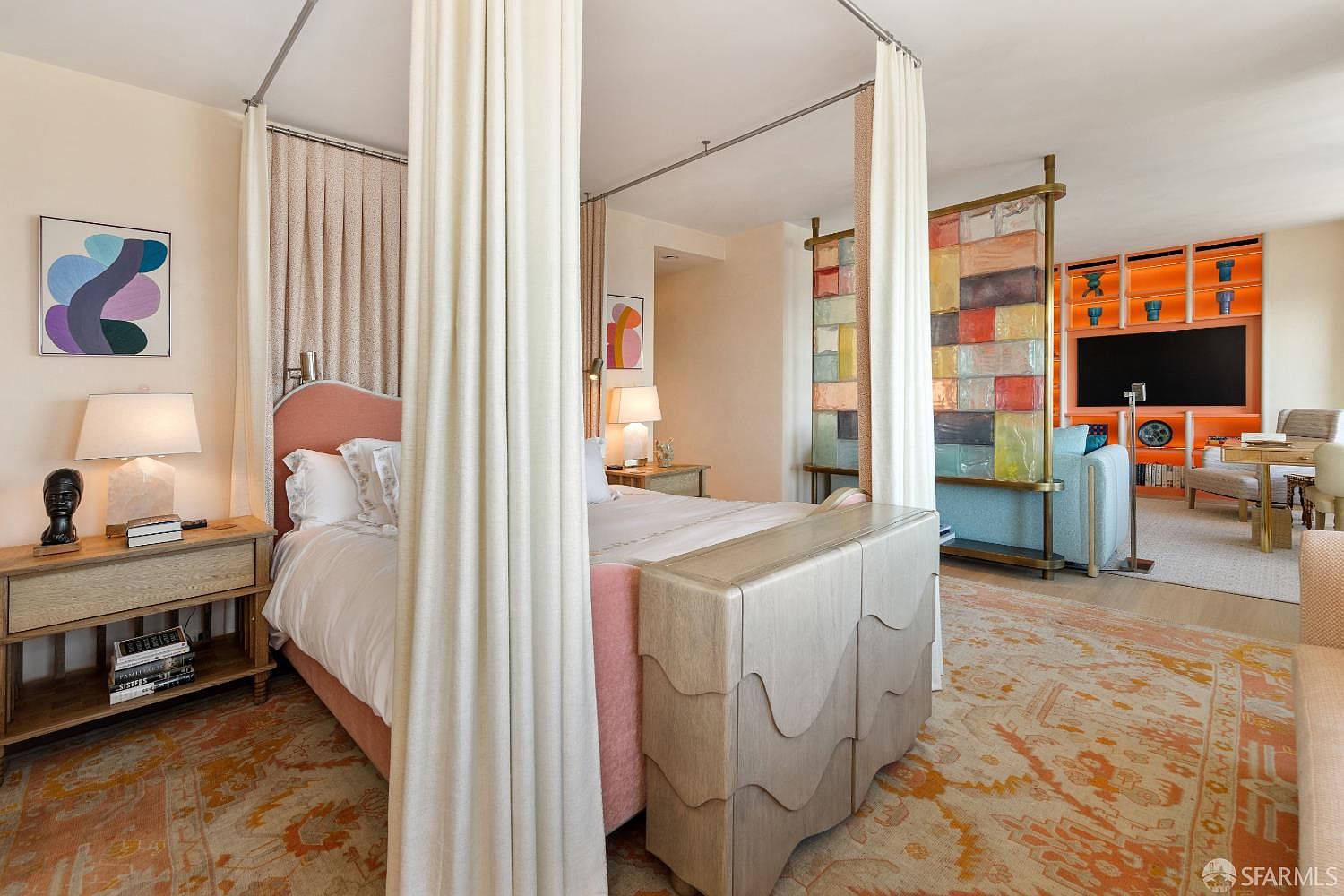This primary bedroom features a distinctive four-poster bed with a soft pink upholstered headboard and white curtains, positioned centrally in the room. A textured, light-toned storage chest sits at the foot of the bed, while a colorful, translucent glass room divider separates the sleeping area from a living space with bright orange shelving. The room is decorated with a patterned area rug and modern art, creating a vibrant and eclectic aesthetic.