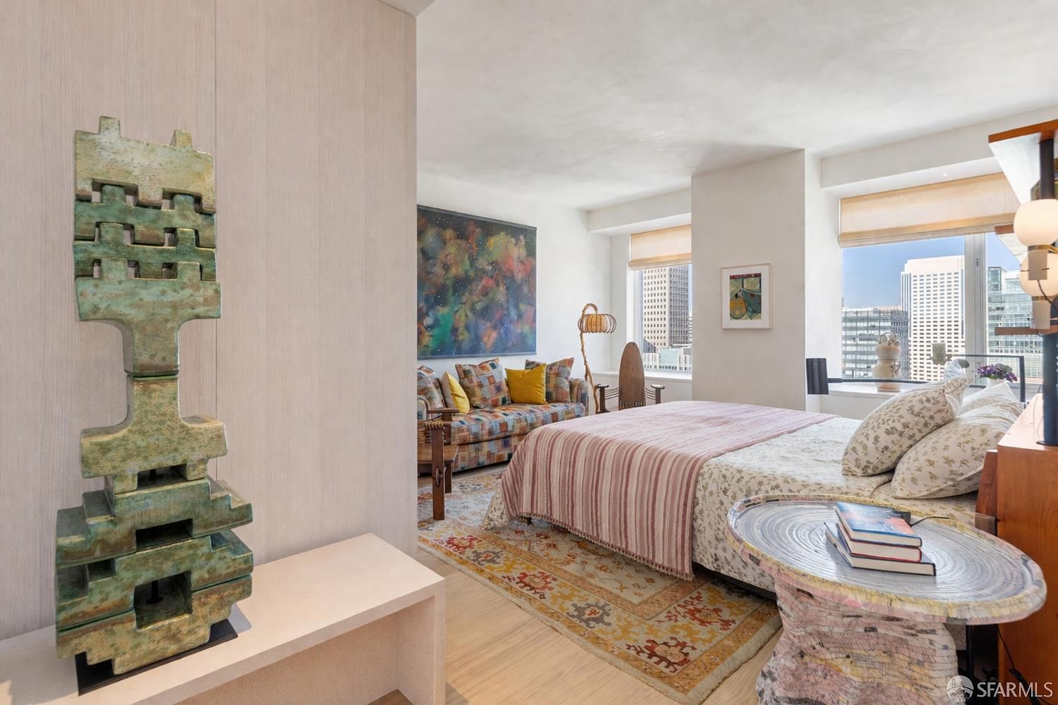 This sophisticated bedroom features a blend of modern and eclectic design, highlighted by a large, abstract piece of art above a patterned sofa. The room includes a bed dressed in light linens with a striped throw, positioned near large windows that offer expansive city views. A unique, textured sculpture stands in the foreground, adding an artistic focal point to the light-filled space.