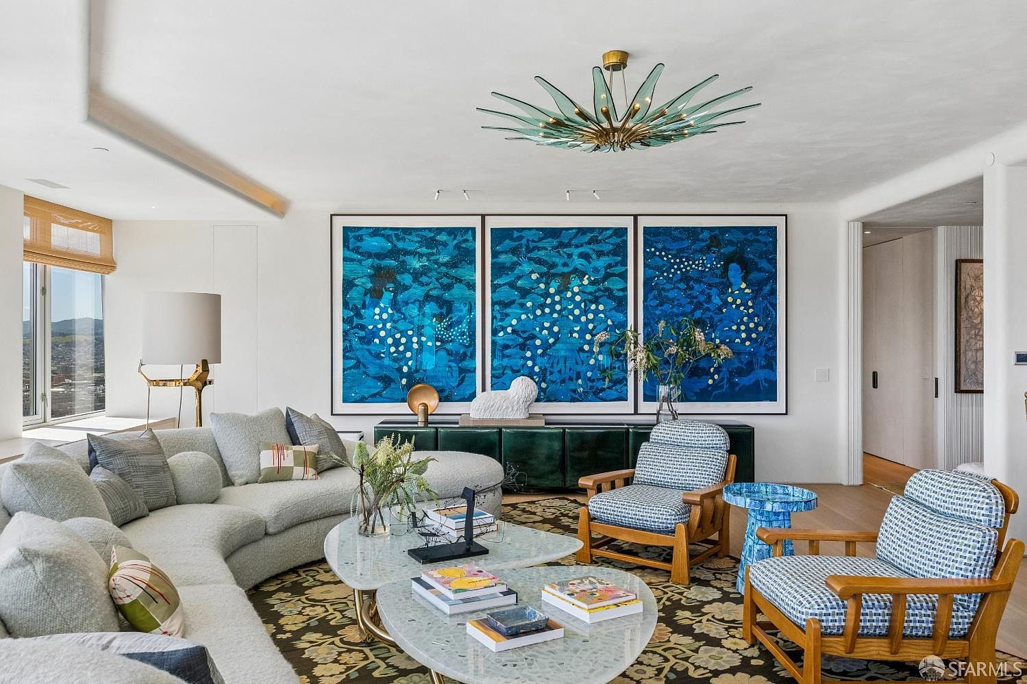 This sophisticated living room features a large, curved light-colored sofa paired with two patterned armchairs and a unique, multi-tiered stone coffee table. The space is anchored by a striking triptych of vibrant blue abstract art hanging above a dark green console table, all illuminated by a dramatic, starburst-style chandelier. Large windows to the left provide ample natural light and a view of the surrounding landscape, creating an elegant and artistic atmosphere.