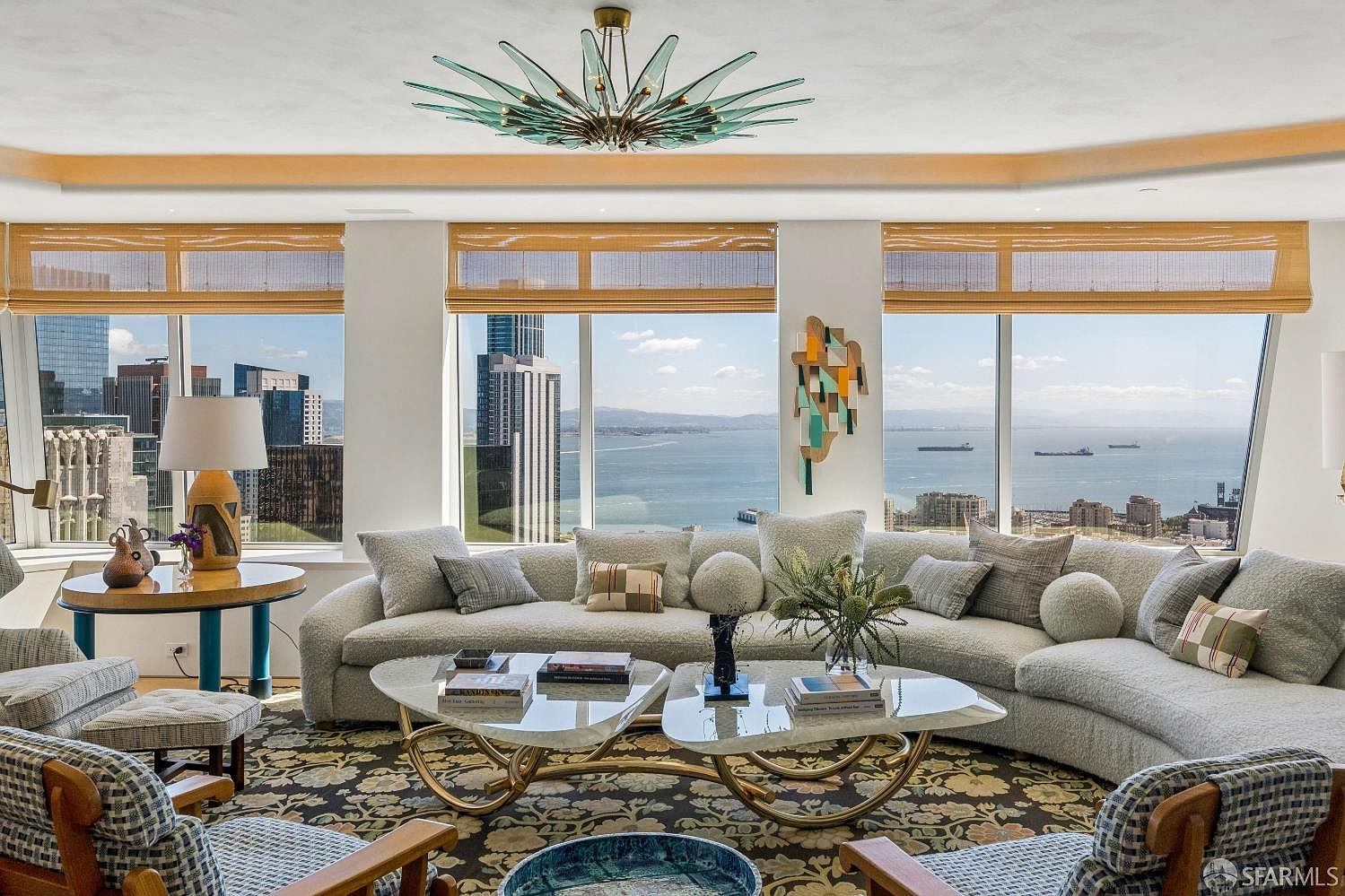 This sophisticated living room features a large, curved sectional sofa arranged around two sculptural, glass-topped coffee tables on a patterned area rug. The space is defined by expansive windows that offer panoramic views of the city skyline and the bay, framed by natural woven shades. A striking, multi-armed glass chandelier serves as a focal point, while the room's neutral color palette and curated decor create an elegant and airy atmosphere.
