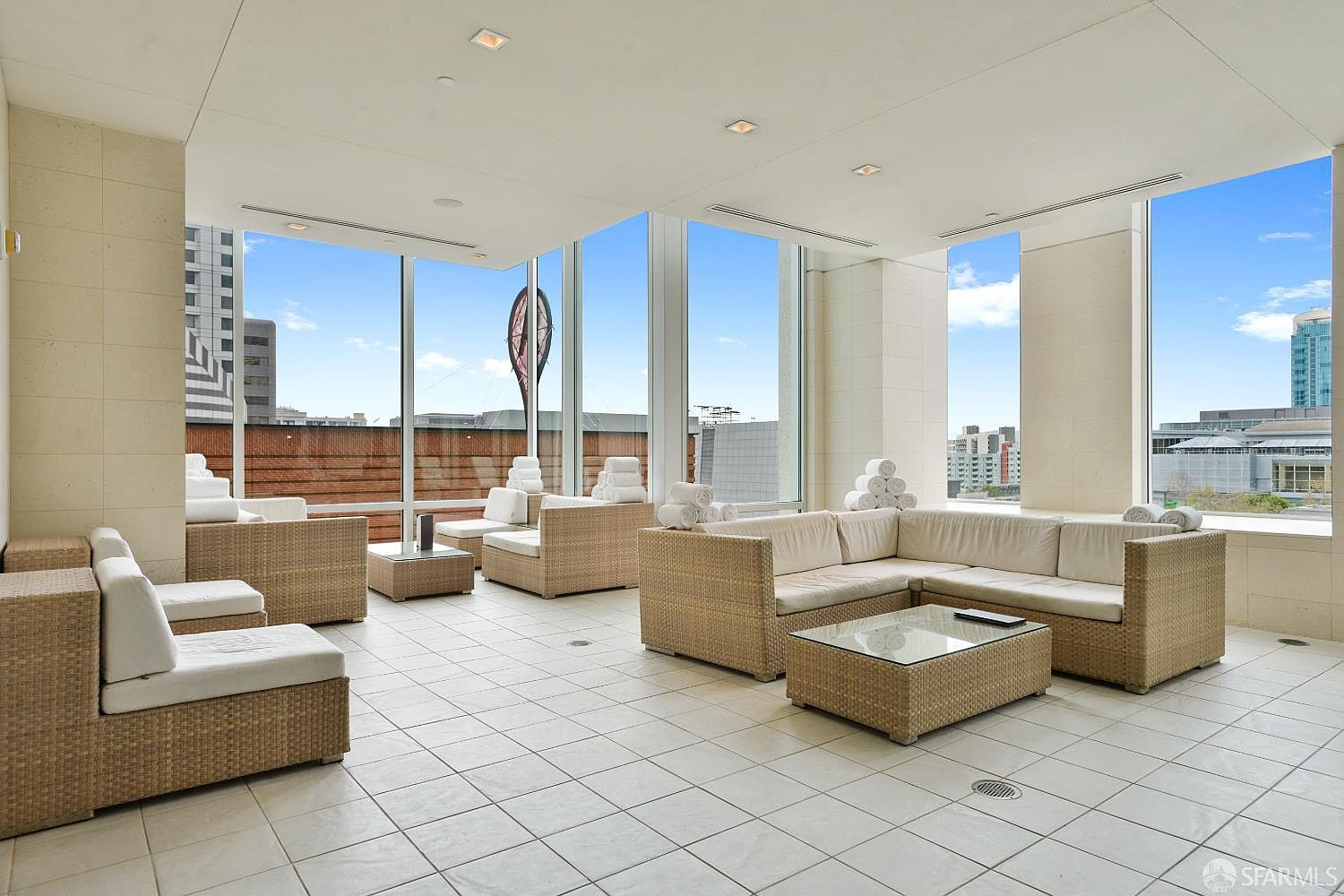 This bright, open-air lounge area features light-colored tile flooring and multiple sets of wicker-style sectional seating with white cushions. Large windows provide expansive views of the surrounding urban landscape, creating a sophisticated and airy atmosphere. The space is designed for relaxation, with stacks of white towels placed on the furniture, suggesting a spa-like or pool-side amenity.