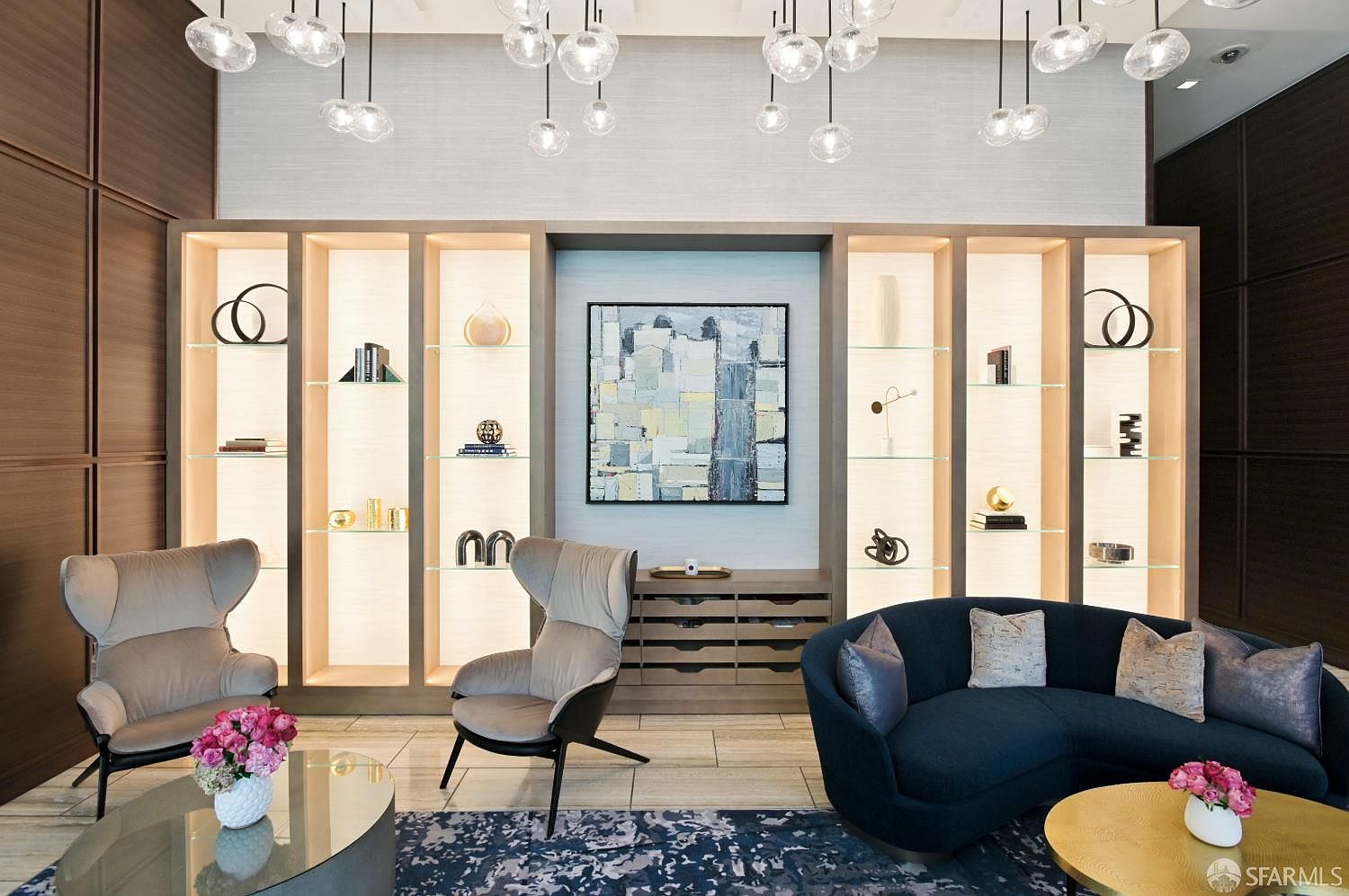 This sophisticated living area features a symmetrical arrangement with two modern armchairs and a curved navy sofa centered around a large, illuminated built-in shelving unit. The wall display showcases curated decor and a central abstract painting, while a cluster of glass pendant lights hangs from above to create a dramatic focal point. The space is grounded by a patterned area rug and polished stone flooring, offering a refined and luxurious atmosphere.