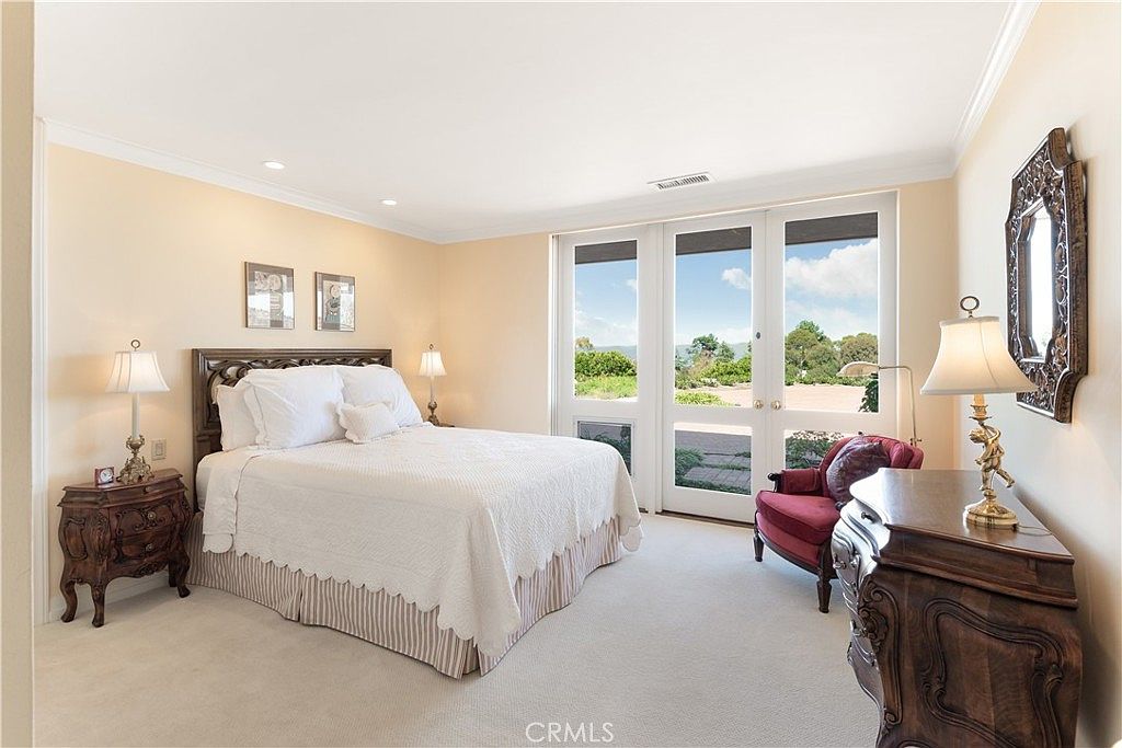 This spacious primary bedroom features a neutral color palette, plush carpeting, and a large bed with a dark wood headboard. Tall windows with glass doors provide ample natural light and a view of the outdoor landscape. The room is furnished with ornate dark wood nightstands and a matching dresser, creating a traditional and elegant atmosphere.