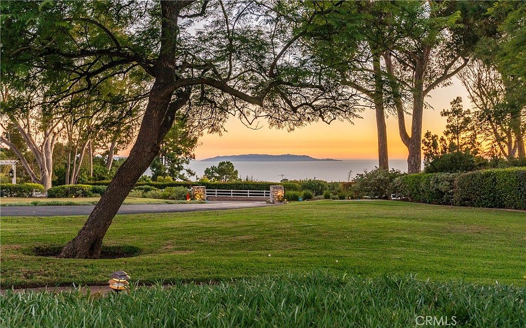 This expansive, well-manicured lawn offers a serene outdoor setting framed by mature trees and lush greenery. The perspective looks out toward a picturesque ocean view with a distant island silhouette against a vibrant sunset sky. A white fence and stone-capped pillars mark the property boundary, creating a peaceful and private atmosphere.