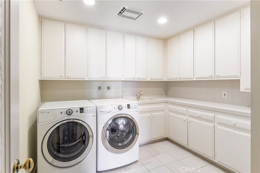 This functional laundry room features white cabinetry providing ample storage space above and below the counter. A side-by-side washer and dryer set is positioned next to a built-in utility sink, all set against a neutral tiled floor. The room is brightly lit by recessed ceiling lights, creating a clean and organized workspace.