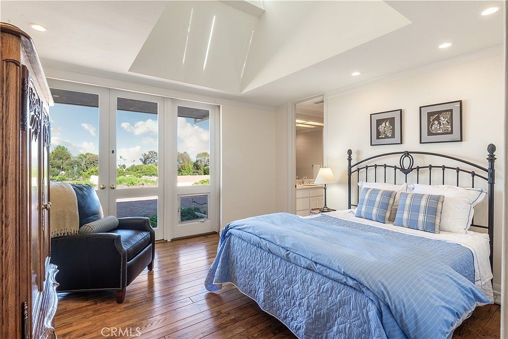 This bright and airy bedroom features a vaulted ceiling with a skylight, hardwood flooring, and a large set of glass doors that open to an outdoor view. The room is furnished with a dark metal-framed bed topped with blue and white bedding, a leather armchair, and a large wooden armoire. The space has a clean, traditional aesthetic with neutral walls and a doorway leading to an adjoining bathroom.