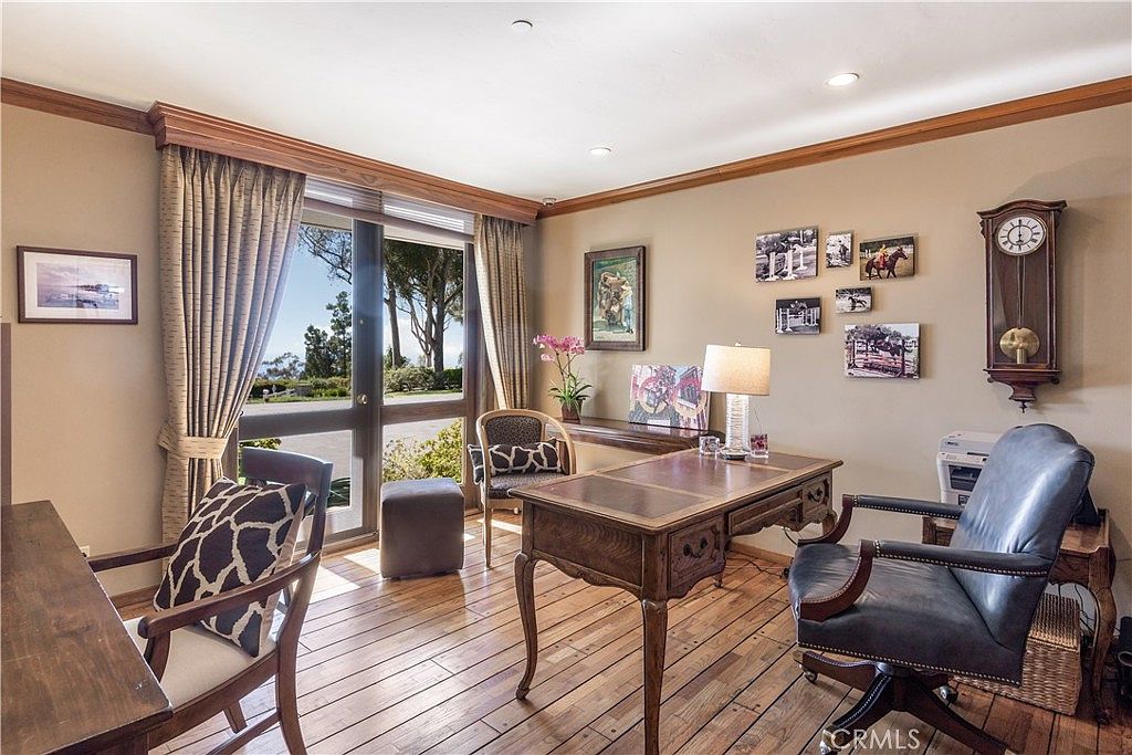 This home office features warm-toned wood flooring and a classic, professional aesthetic with a large central desk and a leather executive chair. A wall of large windows provides ample natural light and a view of the outdoors, while a traditional grandfather clock and a gallery wall of framed photos add character to the space. The room is well-appointed with a side chair and a small console table, creating a functional and inviting workspace.