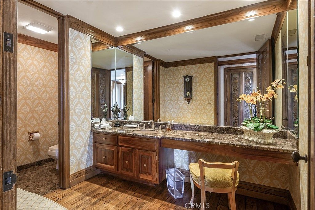 This traditional bathroom features a long vanity with a stone countertop, dark wood cabinetry, and a dedicated makeup seating area. The walls are adorned with patterned wallpaper, complemented by a large mirror and warm recessed lighting. A wooden floor adds a rustic touch to the space, which includes a separate water closet area visible through an open doorway.
