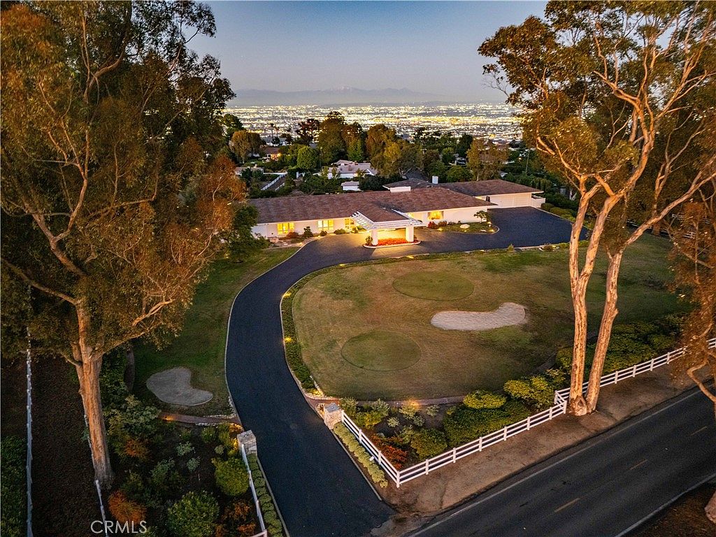 This high-angle aerial view captures a sprawling single-story residence set on a large, manicured lot featuring a private putting green and sand traps. A long, curved asphalt driveway leads from the street to a covered porte-cochère entrance, while the background showcases a panoramic view of a city skyline at dusk. The property is framed by mature trees and a white picket fence, creating a sense of privacy and luxury.