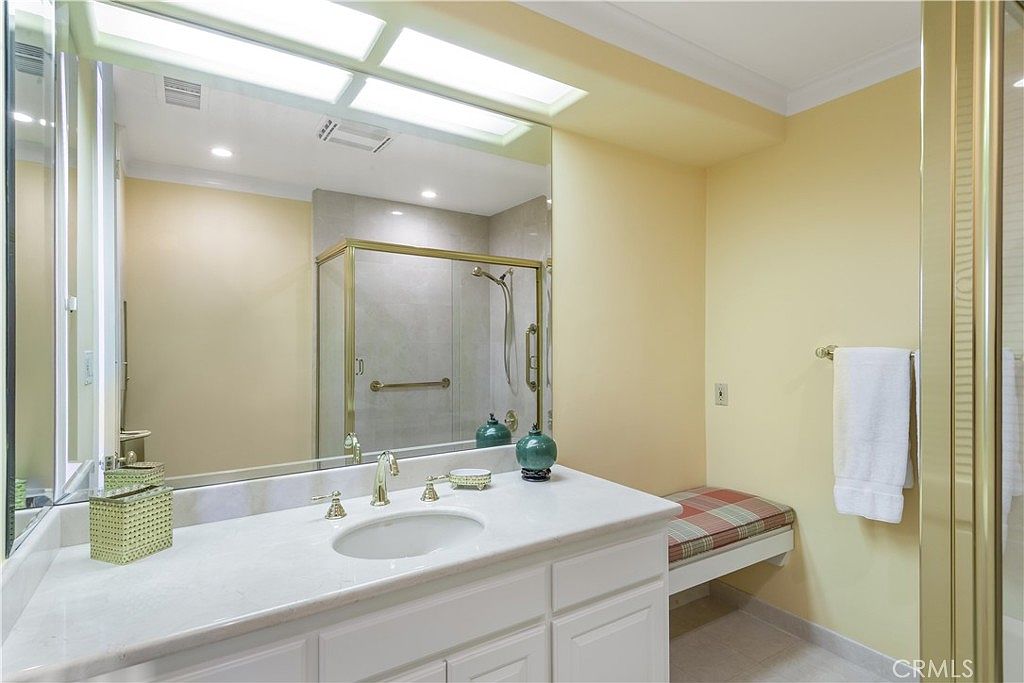 This bathroom features a white vanity with a light-toned stone countertop and a gold-toned faucet. A large, wall-to-wall mirror reflects the shower enclosure, which is framed in gold-toned metal and contains a tiled interior. To the right, a built-in bench with a plaid cushion provides seating, and a white towel hangs on the wall, creating a functional and traditional aesthetic.