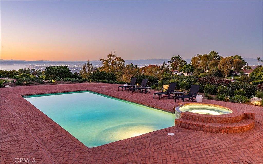 This expansive outdoor space features a large, rectangular swimming pool paired with a circular raised spa, both set within a wide brick-paved patio. Several lounge chairs are arranged on the deck, which overlooks a scenic, elevated view of the surrounding landscape and distant horizon under a soft sunset sky. The setting offers a serene and luxurious atmosphere, perfect for outdoor relaxation and entertaining.