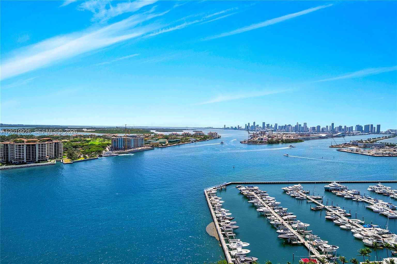 This high-angle aerial view captures a vibrant coastal scene featuring a large marina filled with numerous boats docked in clear blue water. In the distance, a sprawling cityscape skyline rises against a bright blue sky, while a nearby residential complex sits along the lush green shoreline. The perspective provides a comprehensive look at the waterfront lifestyle and the expansive waterway connecting the marina to the urban horizon.