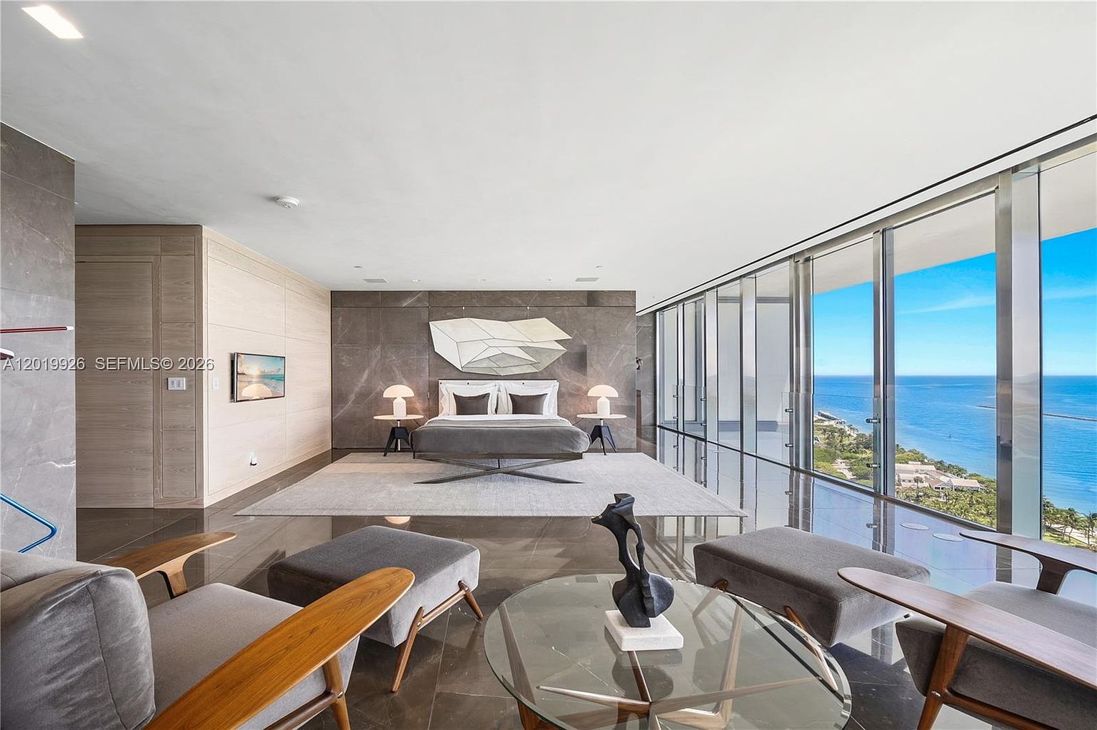 This expansive primary bedroom features a minimalist, modern aesthetic with a neutral color palette and dark stone flooring. The room is anchored by a central bed positioned against a large, dark stone wall feature, while floor-to-ceiling windows provide an unobstructed, panoramic view of the ocean and coastline. In the foreground, a glass-topped coffee table and two armchairs create a sophisticated seating area, emphasizing the luxurious and airy atmosphere of the space.