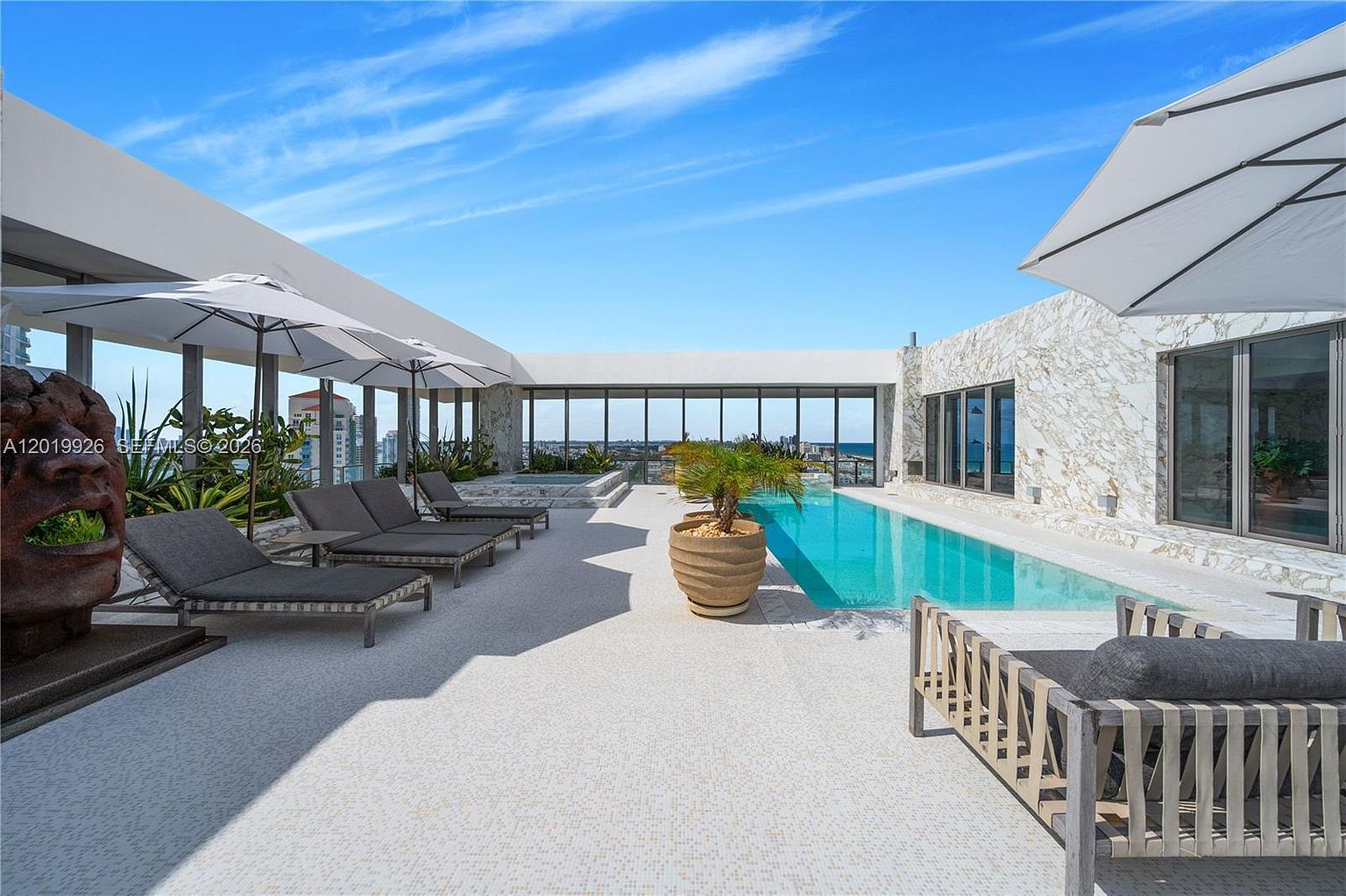 This luxurious rooftop terrace features a sleek, rectangular swimming pool and a separate spa area, set against a backdrop of expansive city and ocean views. The deck is furnished with multiple lounge chairs under umbrellas, creating a sophisticated outdoor retreat. The space is defined by clean architectural lines, stone-clad walls, and a modern, minimalist aesthetic.
