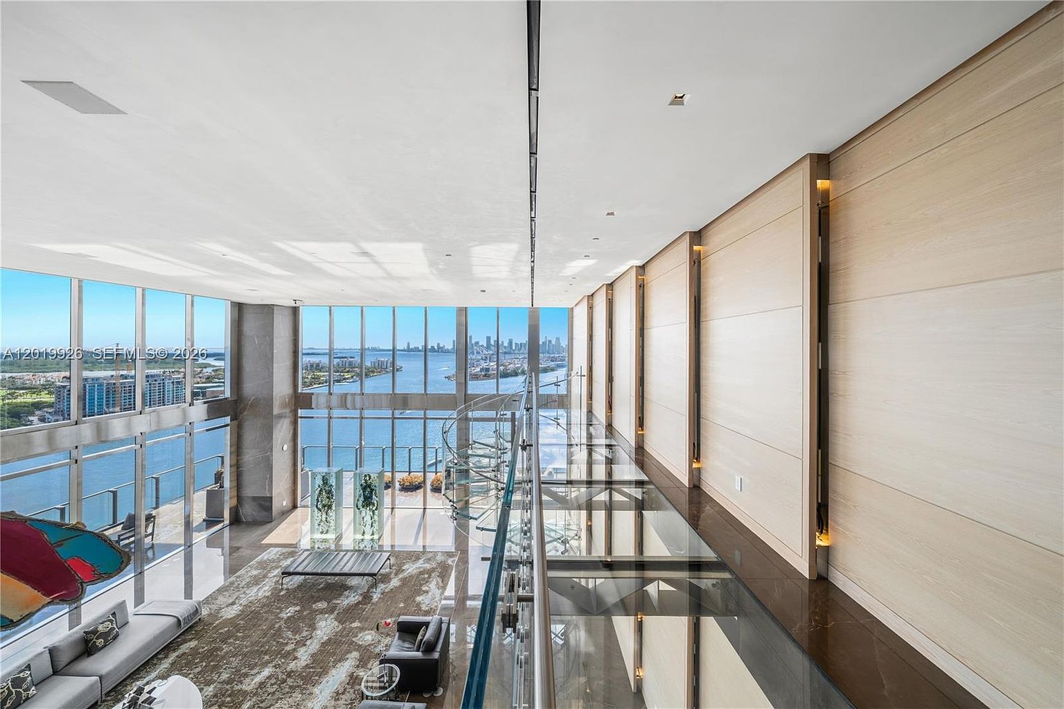 This expansive, double-height living area features a dramatic glass-floored mezzanine overlooking the main floor, which is bathed in natural light from massive floor-to-ceiling windows. The space showcases a sophisticated, modern aesthetic with light wood wall paneling, polished dark flooring, and a sweeping spiral staircase. The panoramic view of the bay and city skyline creates a luxurious, airy atmosphere that emphasizes the property's height and architectural grandeur.