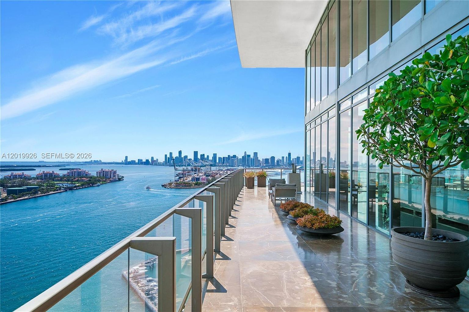 This expansive, high-rise balcony offers a panoramic view of the bay and the distant city skyline. The space features polished stone flooring, a glass railing, and a large potted tree, creating a sophisticated outdoor living area. Floor-to-ceiling glass walls provide a seamless transition from the interior to this impressive vantage point.