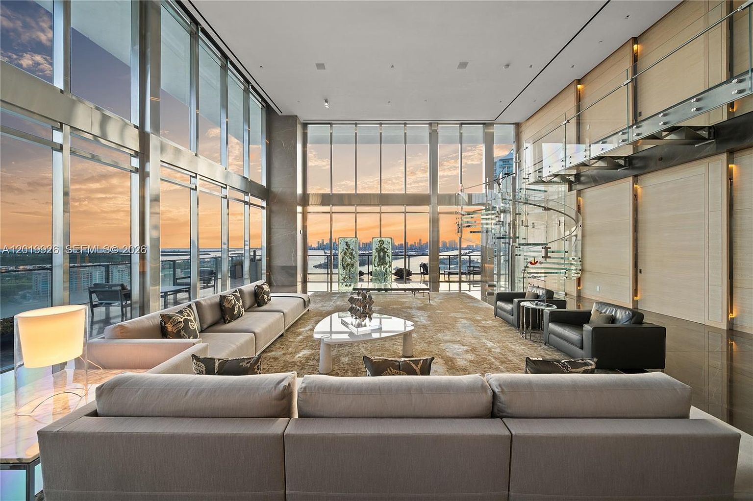 This expansive, double-height living room features dramatic floor-to-ceiling windows that offer panoramic city and water views. The space is anchored by a large, neutral-toned sectional sofa and a modern, sculptural coffee table set upon a textured area rug. A striking glass spiral staircase adds a contemporary architectural element, while the minimalist design emphasizes the vast scale and natural light of the room.