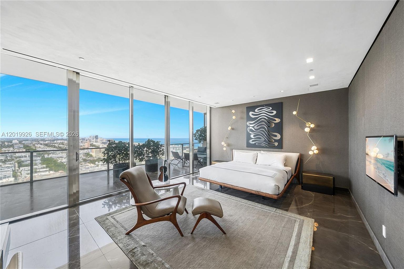This luxurious primary bedroom features a sleek, modern aesthetic with dark, polished flooring and a large area rug. The room is anchored by a platform bed positioned against a dark accent wall adorned with abstract art and unique, branching wall-mounted light fixtures. Expansive floor-to-ceiling windows provide a panoramic view of the city skyline and the ocean, while a comfortable lounge chair and ottoman offer a perfect spot to enjoy the scenery.