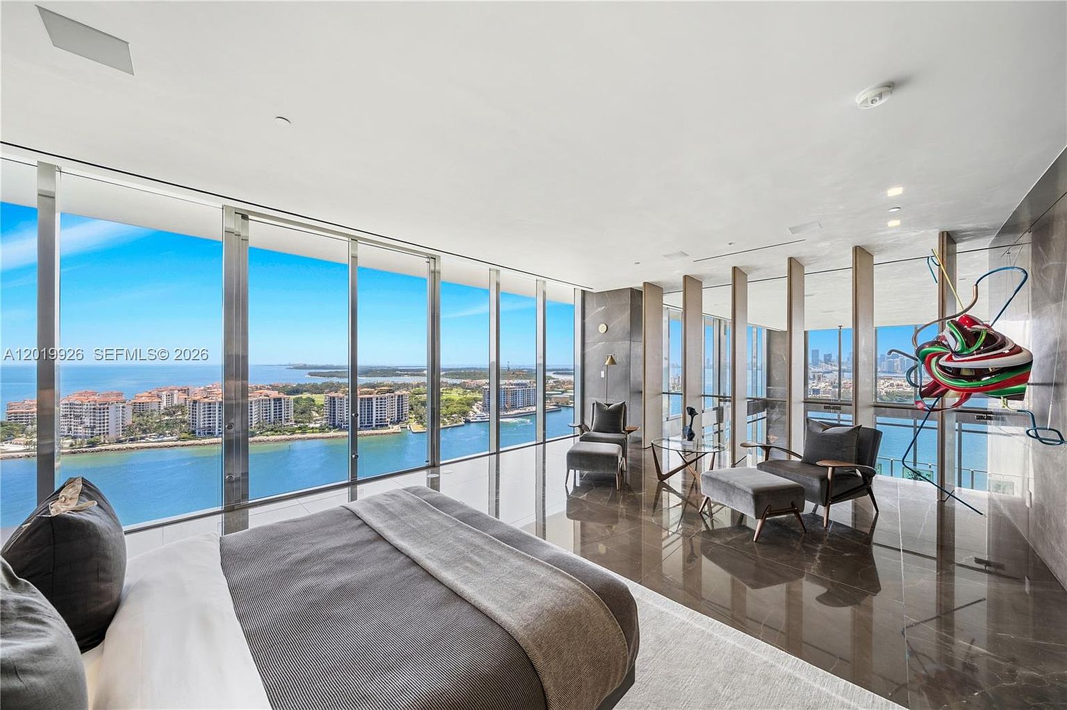 This expansive primary bedroom features floor-to-ceiling windows that offer panoramic views of the bay and city skyline. The room is finished with dark, polished stone flooring and includes a seating area with two armchairs and ottomans, as well as a modern, abstract sculpture mounted on the wall. The perspective is wide-angle, emphasizing the seamless transition between the luxurious interior and the vast outdoor scenery.