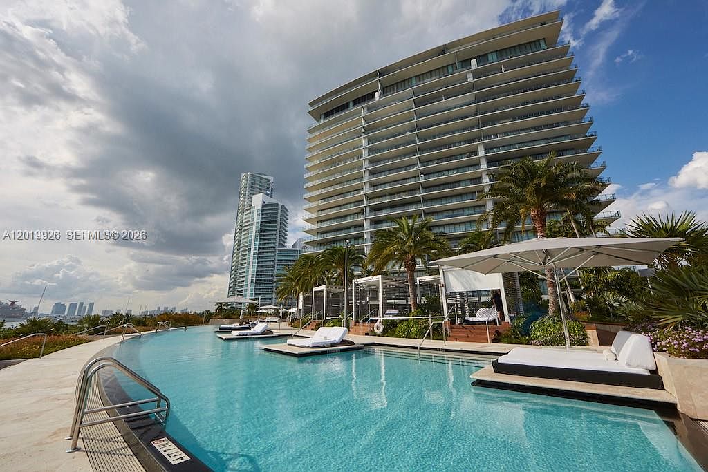 This image captures a luxurious infinity-edge swimming pool at a high-rise residential complex, offering a serene atmosphere with lounge chairs and cabanas. The perspective is from the pool deck, looking toward the modern, multi-story building with its expansive glass balconies and the surrounding urban skyline. The scene conveys a sophisticated, resort-style living experience with clear blue skies and lush palm trees.