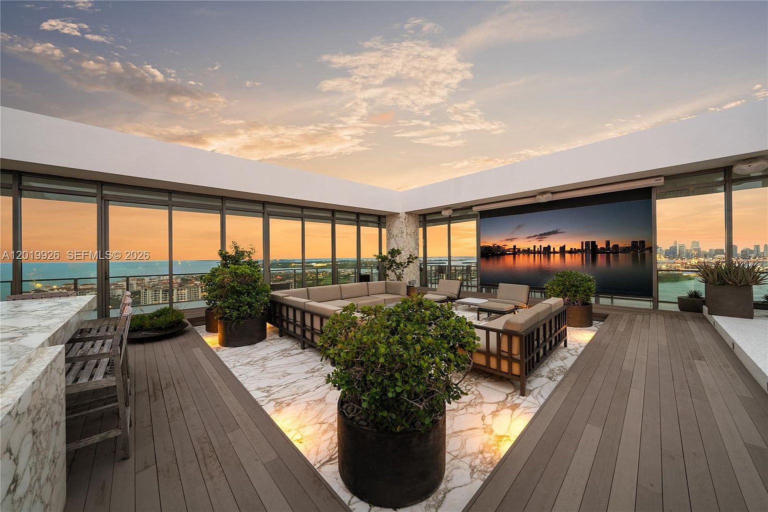 This expansive rooftop terrace offers a luxurious outdoor living space with panoramic city and water views. The area features a large sectional sofa arranged on a patterned stone floor, surrounded by potted greenery and framed by floor-to-ceiling glass walls. A retractable projection screen is positioned to create an open-air cinema experience, while the warm sunset lighting enhances the sophisticated and tranquil atmosphere.