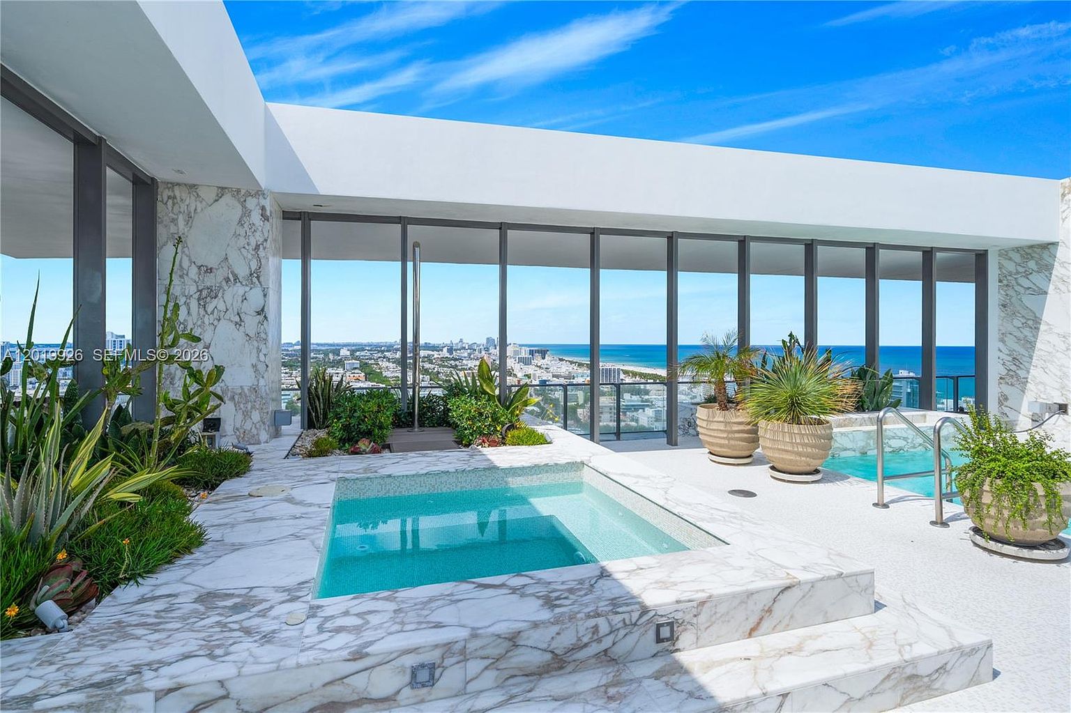 This luxurious rooftop terrace features a sleek, stone-clad spa pool as its centerpiece, set against a backdrop of expansive floor-to-ceiling glass walls that offer panoramic ocean and city views. The area is accented with lush potted greenery and a second, smaller pool, creating a serene and high-end outdoor retreat. The bright, natural lighting and clean architectural lines emphasize a sophisticated, modern aesthetic perfect for relaxation.