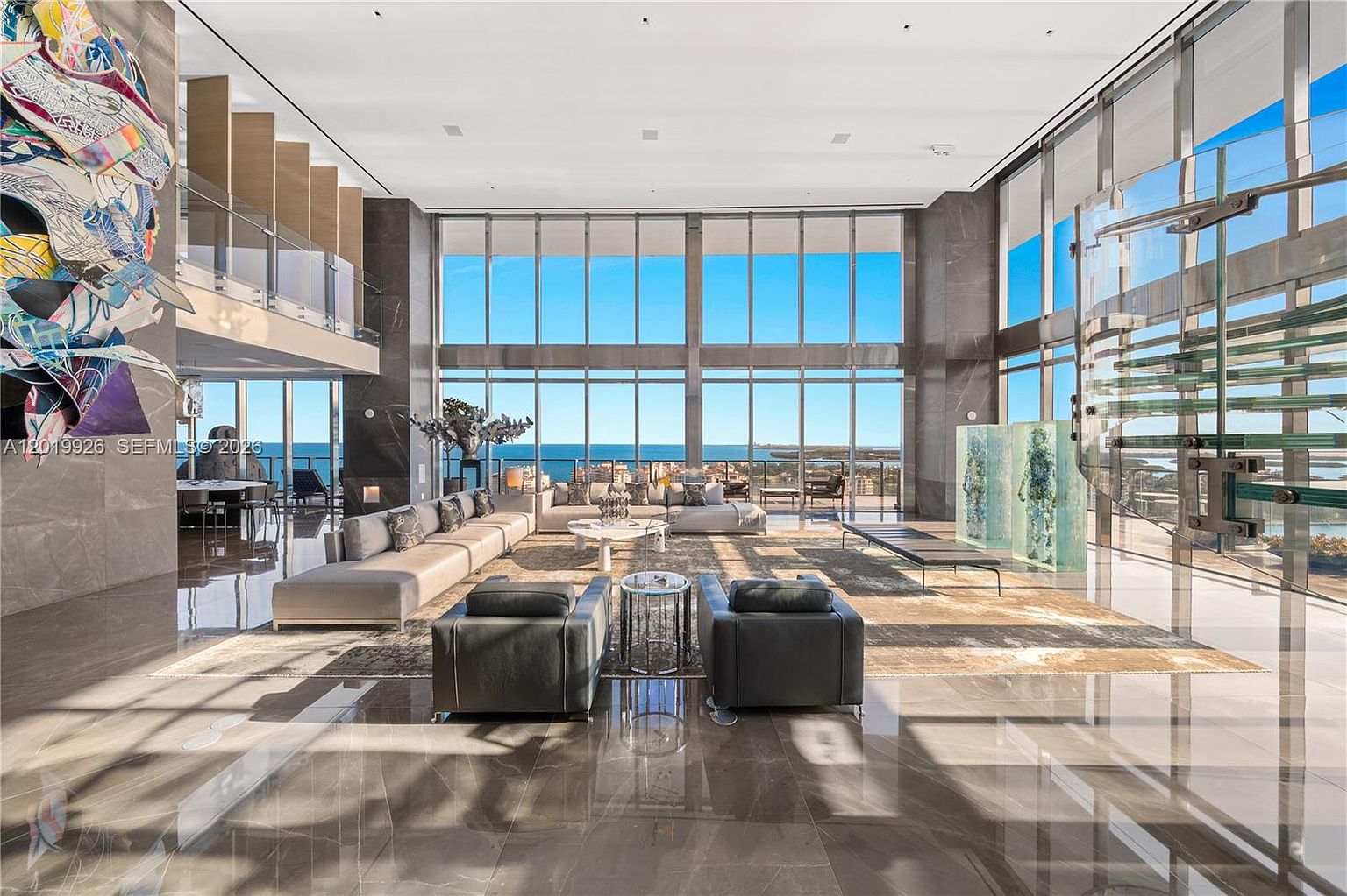 This expansive, double-height living room features a sophisticated open-concept layout with polished stone flooring and massive floor-to-ceiling windows that frame a panoramic ocean view. The space is furnished with a large, modular sectional sofa, two dark leather armchairs, and a central coffee table, all arranged on a textured area rug. A striking, colorful abstract art piece adorns the wall, while a glass-railed staircase and unique sculptural elements add a modern, gallery-like aesthetic to the room.