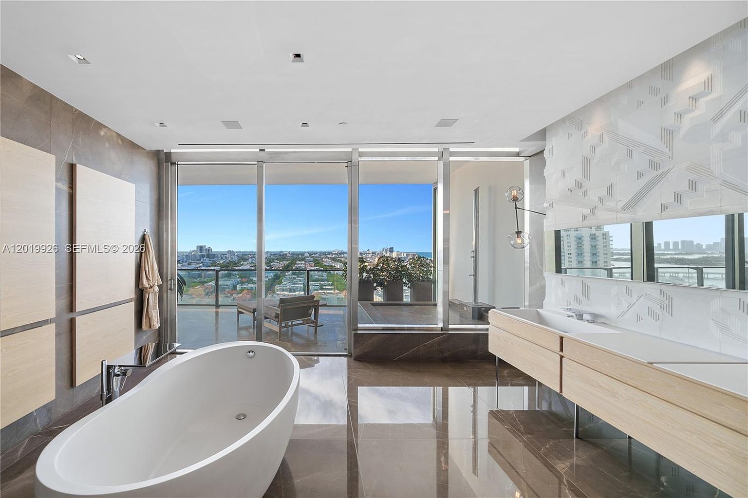 This luxurious primary bathroom features a sleek, freestanding oval soaking tub positioned to overlook a private balcony with expansive city and water views. The space is defined by a floating light wood vanity with a long integrated sink and a textured, geometric-patterned feature wall behind the mirror. Large glass sliding doors provide seamless indoor-outdoor flow, bathing the room in natural light and highlighting the polished stone flooring.