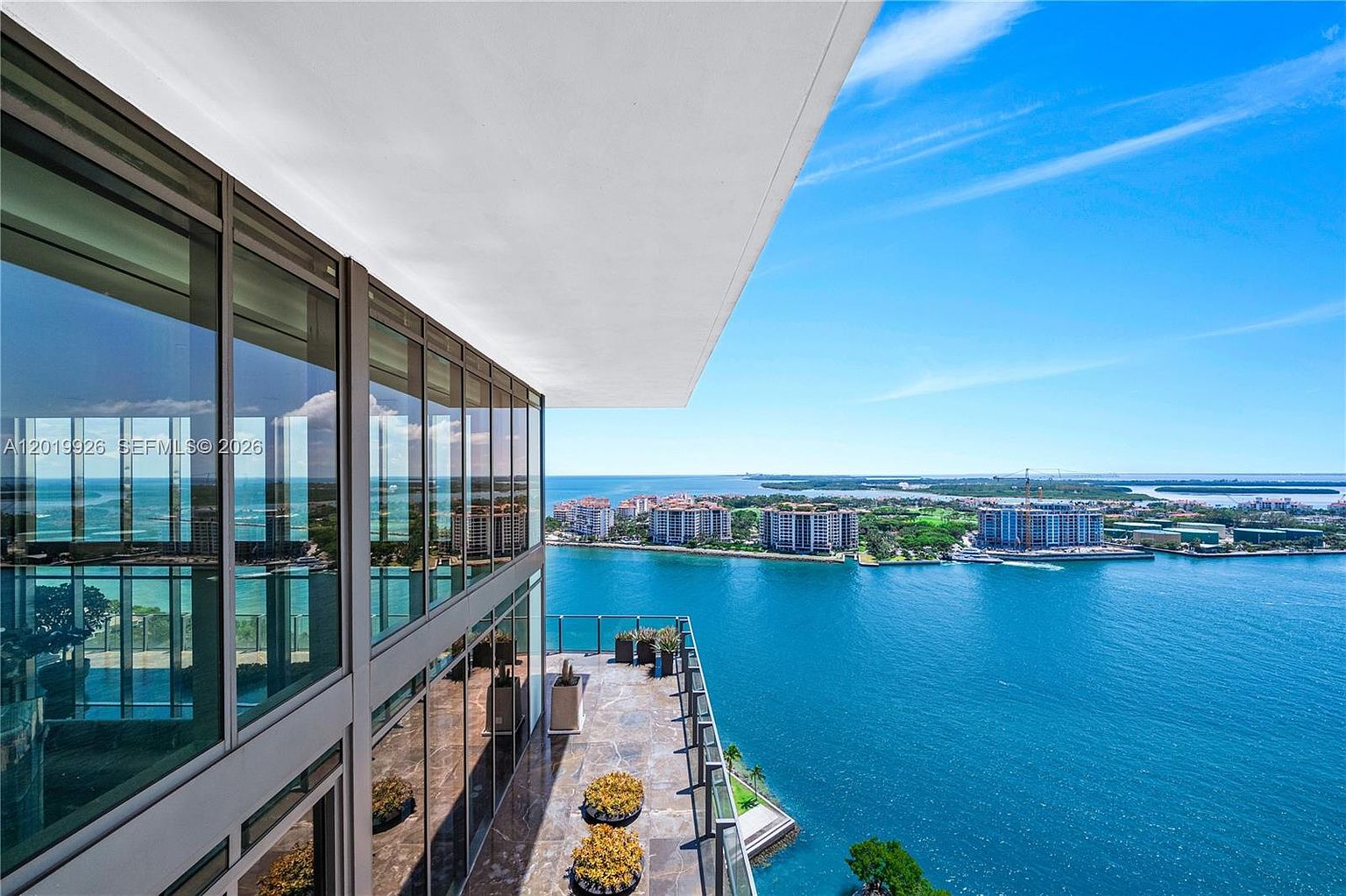 This high-rise balcony offers a sweeping, panoramic view of the bay and distant coastal architecture. The space features stone flooring, potted plants, and a glass railing that maximizes the unobstructed water vistas. Large, floor-to-ceiling windows provide a seamless transition from the interior living space to the outdoor terrace.