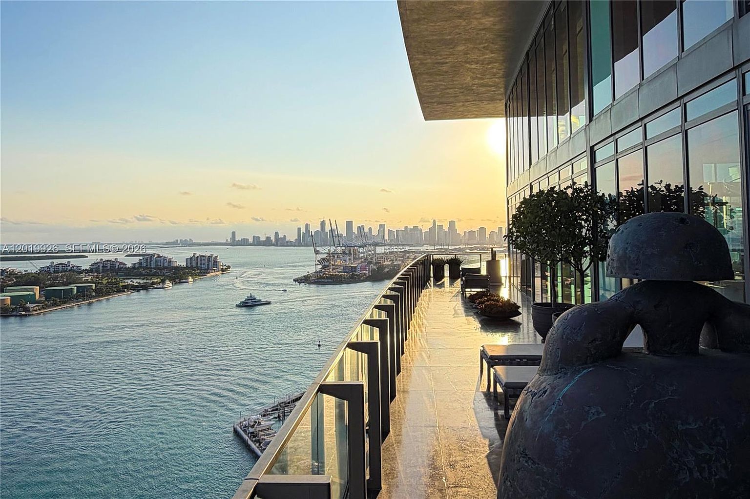 This expansive, high-rise balcony offers a panoramic view of a coastal waterway and a distant city skyline at sunset. The space features a sleek glass railing, a large sculptural art piece in the foreground, and comfortable outdoor seating arrangements along the building's glass facade. The warm, golden light of the setting sun creates a serene and luxurious atmosphere, highlighting the seamless transition between indoor and outdoor living.