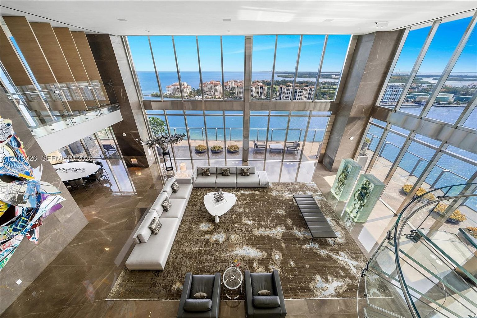 This expansive, double-height living room features dramatic floor-to-ceiling windows that offer panoramic waterfront views. The space is anchored by a large, neutral-toned sectional sofa and a patterned area rug, complemented by a sculptural white coffee table and two dark leather armchairs. A striking, multi-colored abstract art installation hangs near the staircase, while a glass spiral staircase adds a modern architectural element to the bright, open-concept interior.