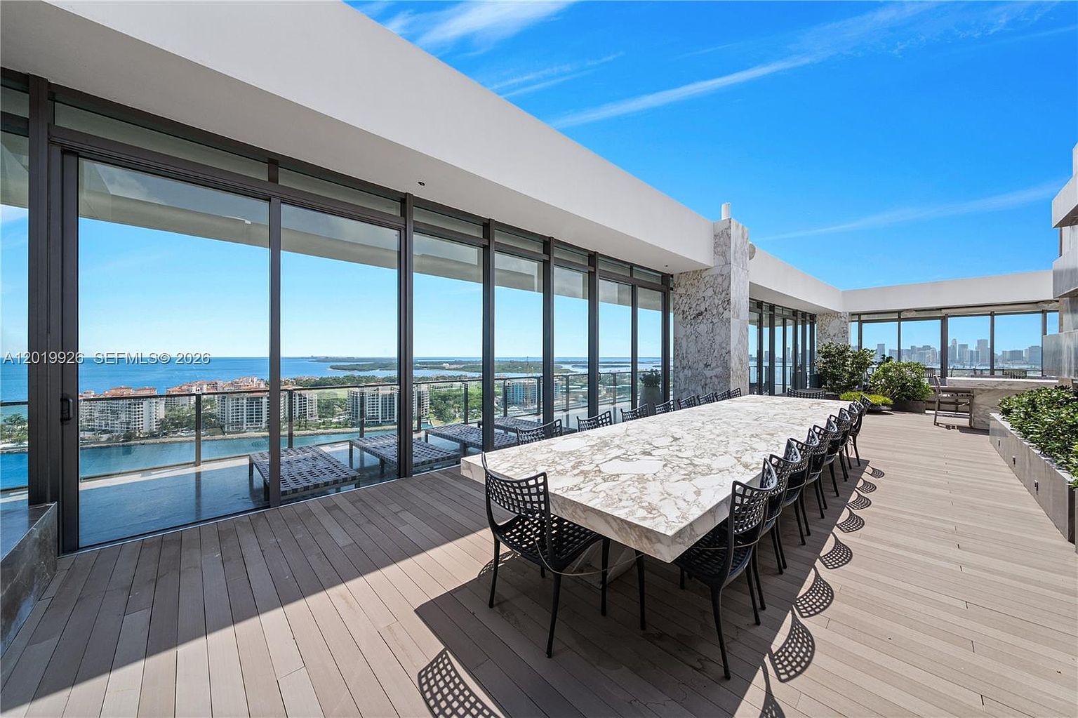 This expansive outdoor terrace features a long dining table with a stone-patterned top, surrounded by modern black metal chairs. The space is enclosed by floor-to-ceiling glass walls that offer panoramic views of the bay and city skyline. The deck is finished with light-toned wood-look planks, creating a sleek and sophisticated atmosphere perfect for outdoor entertaining.