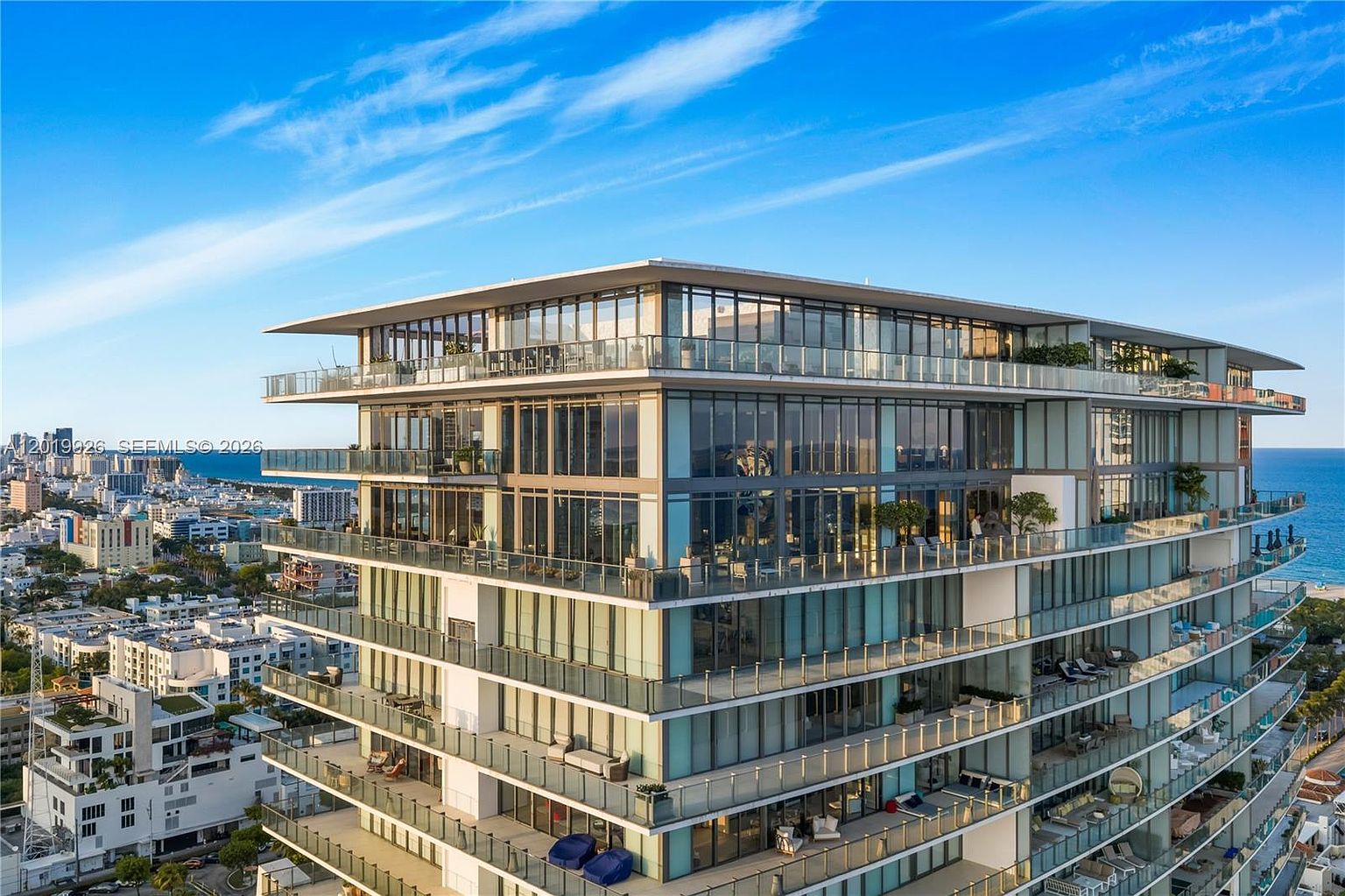 This high-angle aerial shot captures a modern, multi-story luxury condominium building situated along a coastline. The structure features extensive glass facades and expansive, wrap-around balconies on every level, offering panoramic views of the ocean and the surrounding urban landscape. The bright, sunny atmosphere highlights the sleek architectural design and the seamless integration of indoor and outdoor living spaces.
