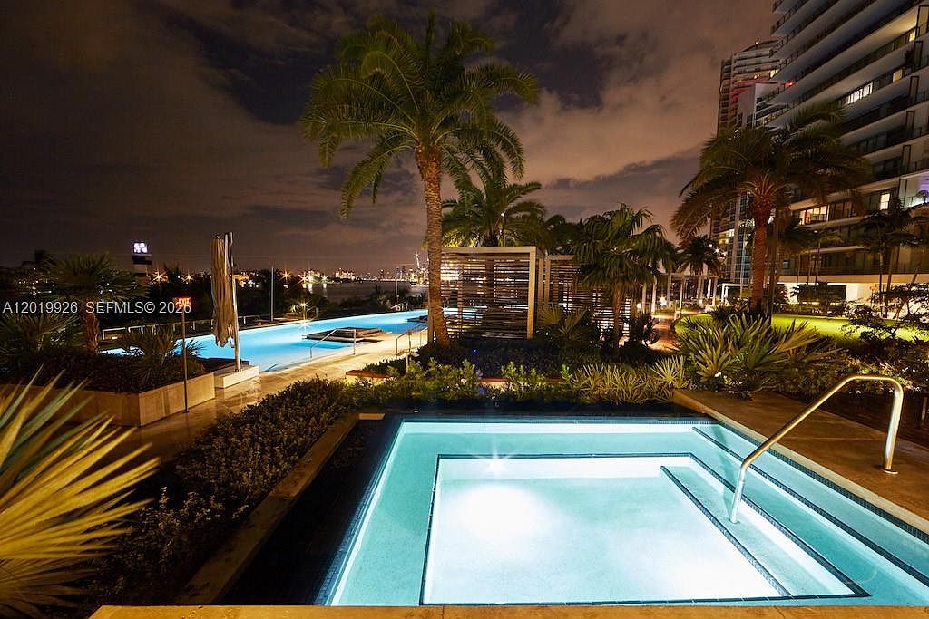 This nighttime shot captures a luxurious resort-style pool and spa area, featuring a glowing, illuminated hot tub in the foreground and a long, sleek lap pool stretching toward the horizon. Lush tropical landscaping, including tall palm trees and manicured shrubs, frames the water features, while a modern high-rise building stands to the right. The scene is set against a dark, cloudy night sky with the distant city lights providing a sophisticated and serene backdrop.