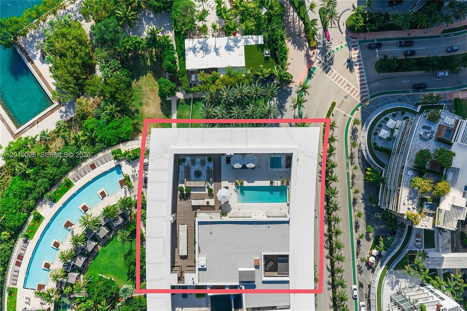 This high-angle aerial view captures a modern residential building rooftop featuring a private terrace with a swimming pool, lounge seating, and sun umbrellas. The surrounding landscape includes lush green trees, manicured lawns, and a winding pathway, with a larger communal pool visible to the left. The perspective provides a clear layout of the building's outdoor amenities and its integration into the surrounding coastal or resort-style environment.