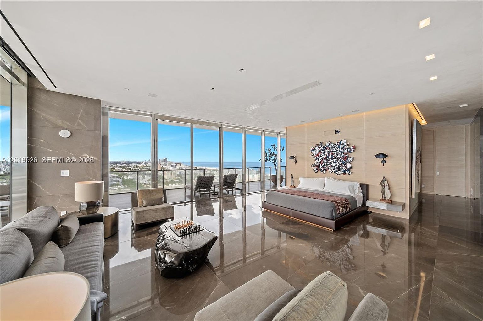 This expansive primary bedroom features a sophisticated open-concept layout with polished stone flooring and floor-to-ceiling windows that provide panoramic ocean and city views. The room is furnished with a low-profile bed, a unique organic-shaped coffee table, and a seating area, all set against a light wood-paneled accent wall adorned with modern art. The overall aesthetic is ultra-luxurious and contemporary, emphasizing a seamless connection between the interior living space and the outdoor horizon.