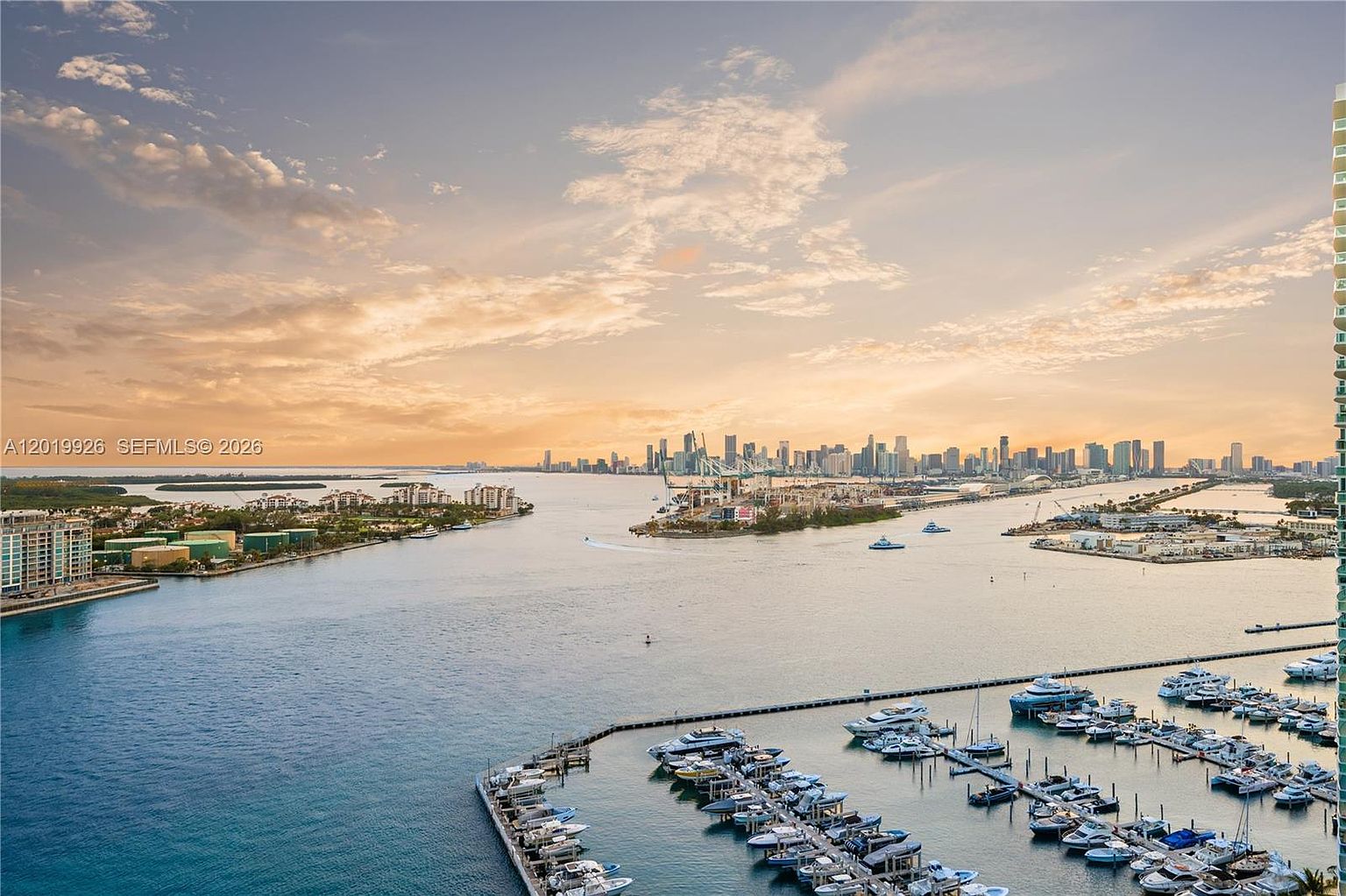 This high-angle aerial view captures a stunning panoramic vista of a coastal waterway, marina, and distant city skyline under a soft, golden-hued sunset. The foreground features a bustling marina filled with numerous yachts and boats, while the mid-ground showcases a wide channel leading toward a busy port area. The composition offers a cinematic perspective, emphasizing the expansive waterfront lifestyle and the vibrant urban backdrop.