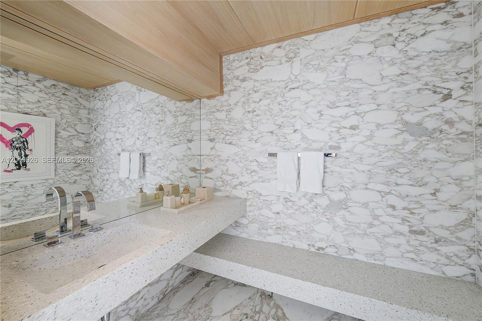 This modern bathroom features a floating vanity and a matching lower shelf crafted from speckled stone, set against a wall of large-format patterned stone tiles. A wide mirror reflects the space, which is accented by light wood ceiling paneling and a framed piece of art on the left wall. The clean lines and neutral color palette create a sophisticated and minimalist aesthetic.