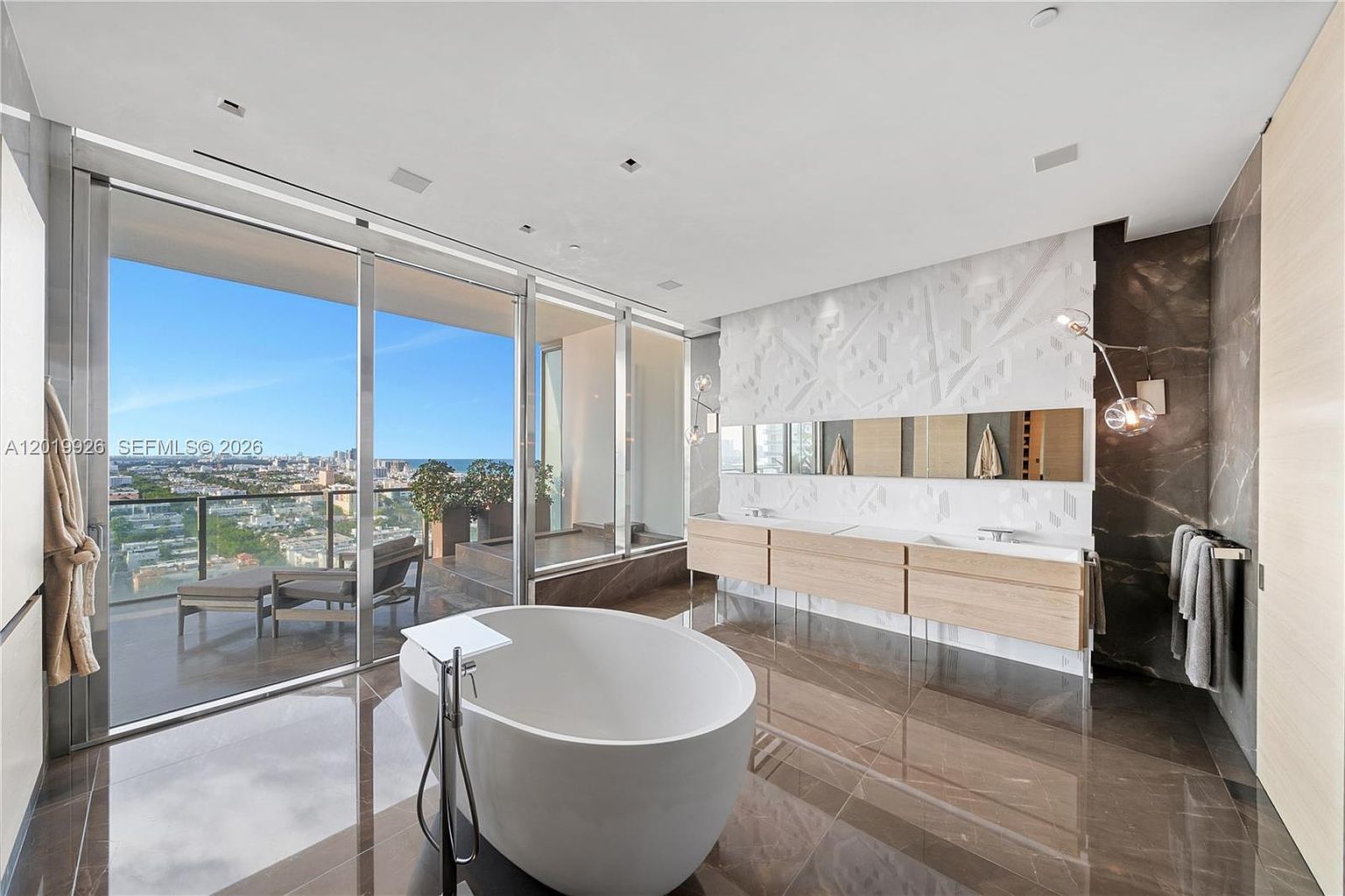 This luxurious primary bathroom features a sleek, freestanding white soaking tub positioned to overlook a private balcony with expansive city and ocean views. The space is defined by dark, polished stone flooring and a long, light wood vanity with dual sinks set against a textured, geometric-patterned wall. Floor-to-ceiling glass panels seamlessly connect the indoor sanctuary to the outdoor terrace, creating an airy and sophisticated atmosphere.