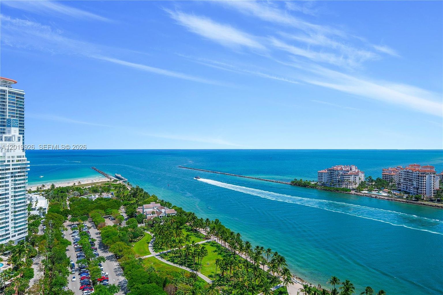 This high-angle aerial view captures a stunning coastal landscape featuring a deep blue ocean inlet, a sandy beach, and a lush green park with palm trees. A tall residential high-rise stands prominently on the left, while a boat leaves a long wake as it travels through the channel toward the open sea. The scene offers a panoramic perspective of a prime waterfront location, emphasizing the proximity to both urban amenities and natural beauty.