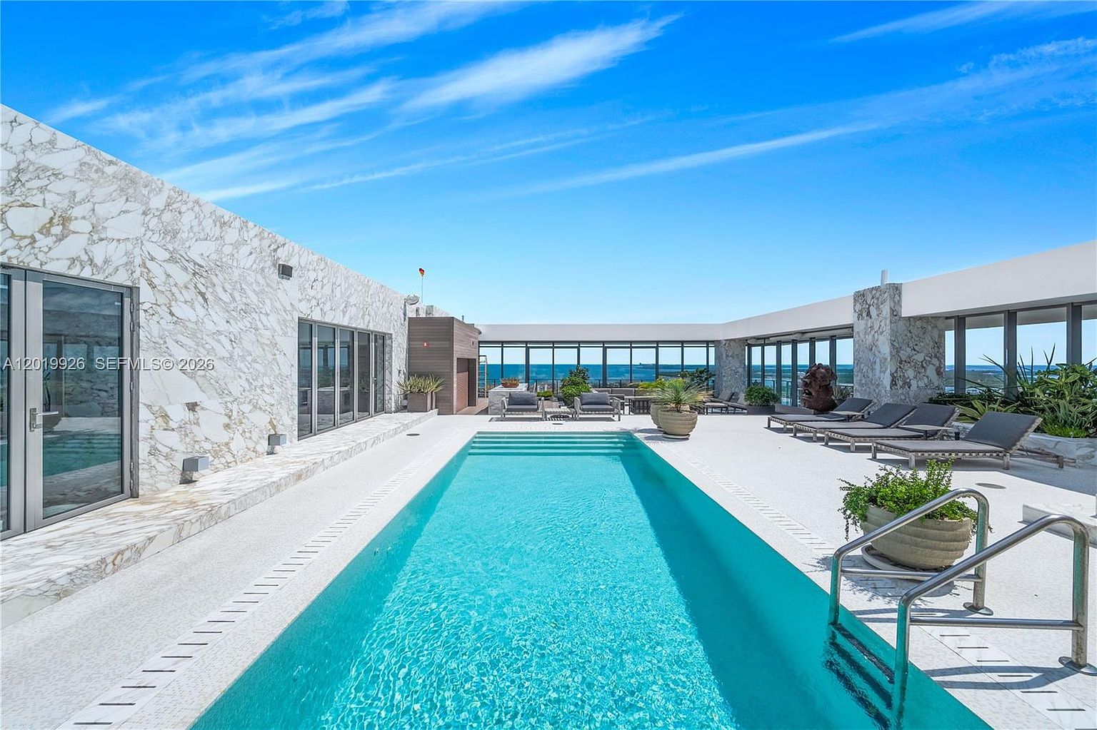 This expansive rooftop terrace features a long, rectangular swimming pool as its centerpiece, set against a backdrop of clear blue skies and distant ocean views. The surrounding deck is finished in light-colored stone and is furnished with multiple lounge chairs, creating a luxurious outdoor retreat. The space is framed by walls clad in white veined stone and large glass panels, emphasizing a modern, high-end architectural aesthetic.