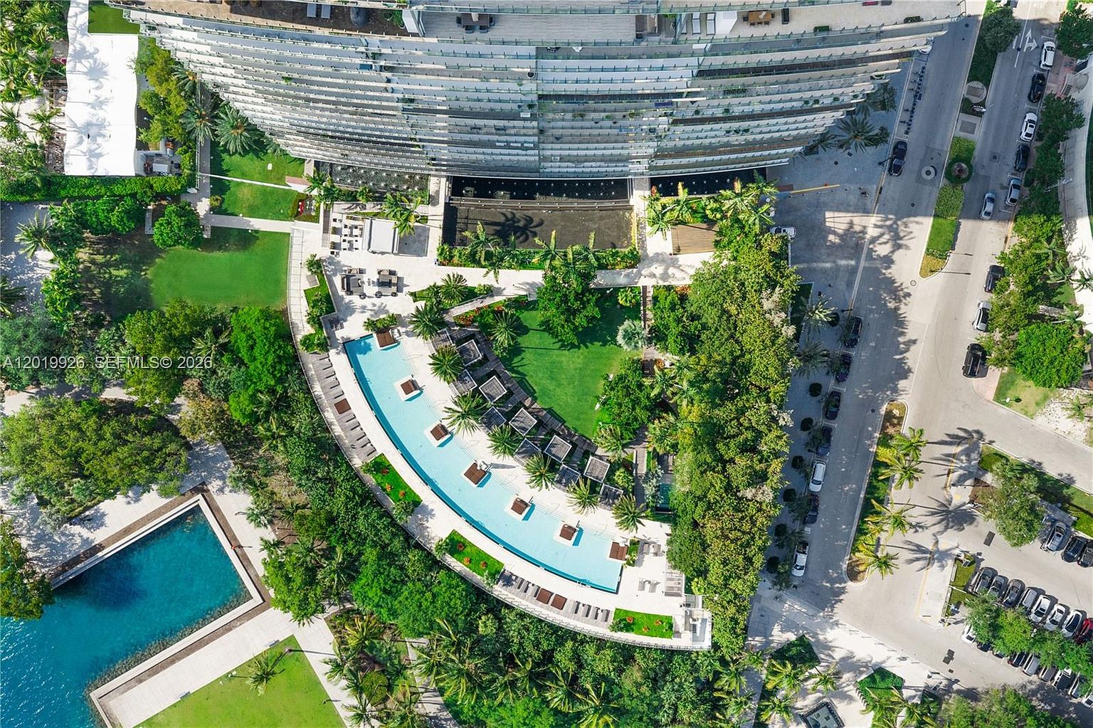 This high-angle aerial view captures a luxury residential complex featuring a curved, multi-story building with extensive glass balconies. The grounds include a large, crescent-shaped swimming pool surrounded by lounge chairs, lush tropical landscaping, and manicured green lawns. A paved road runs along the side of the property, providing access to the building and adjacent parking areas.