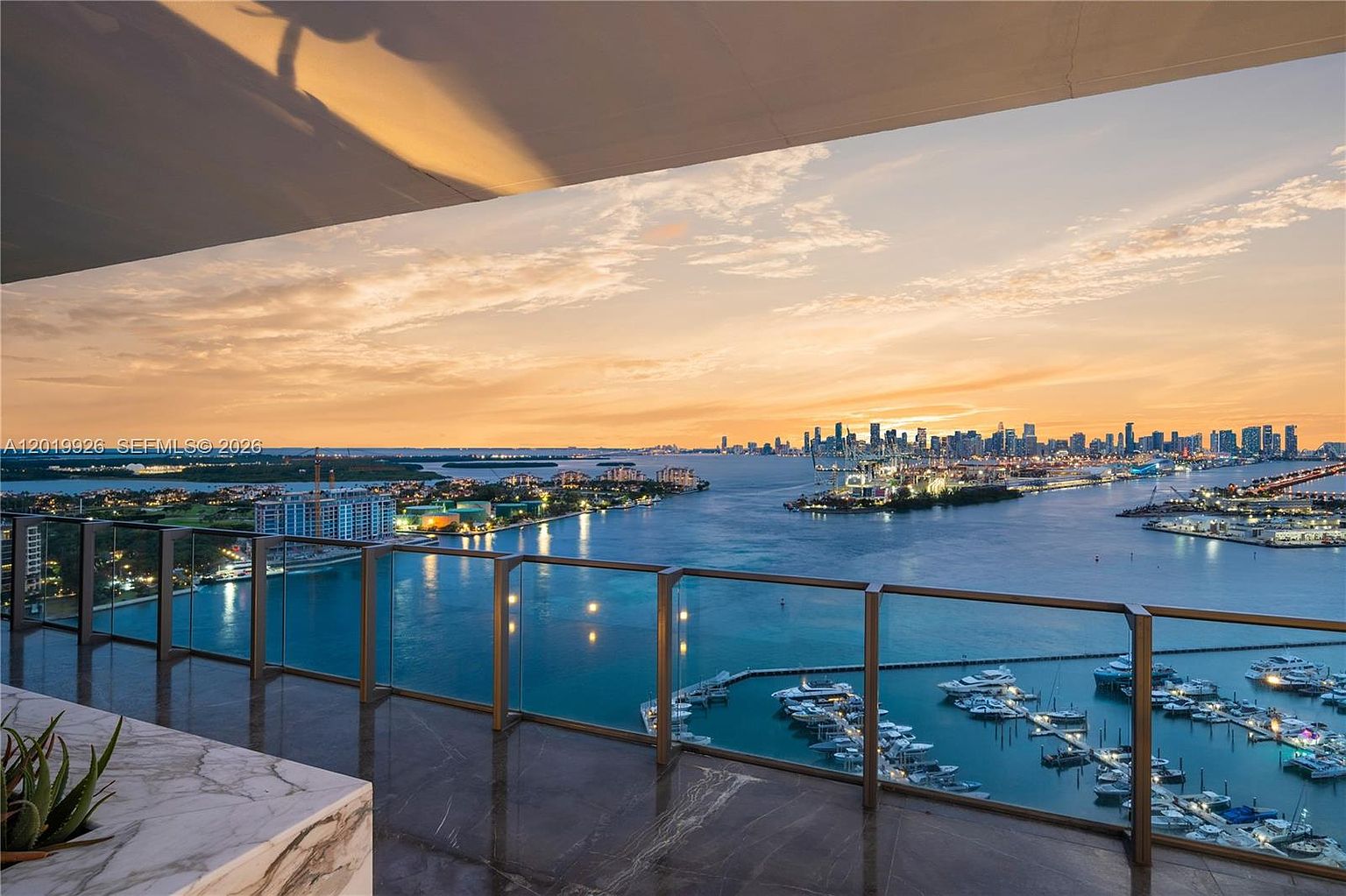 This high-rise balcony offers a sweeping, panoramic view of a harbor and distant city skyline at sunset. The space features a polished stone floor and a sleek glass railing, providing an unobstructed vantage point over the marina below. The warm, golden light of the setting sun reflects off the water, creating a serene and luxurious atmosphere.