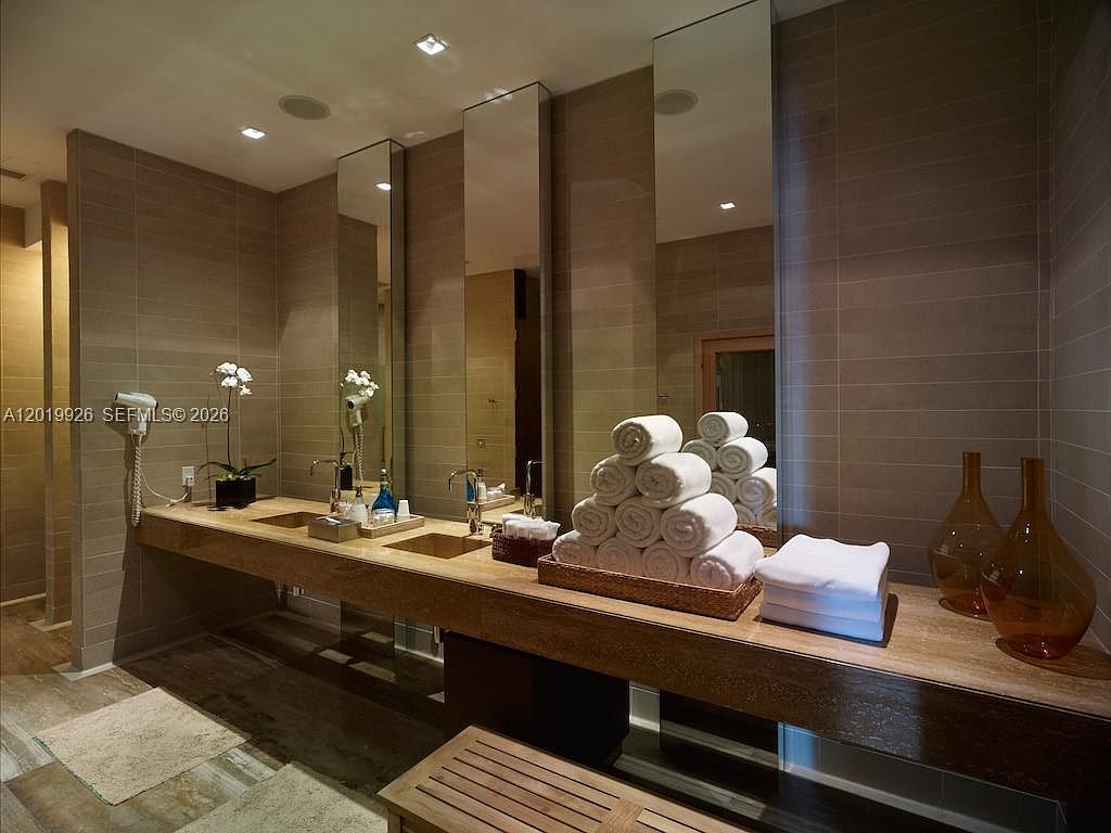 This sophisticated bathroom features a long, floating vanity with a stone countertop and dual integrated sinks, set against a wall of large, neutral-toned tiles. Tall, vertical mirrors are positioned above each sink, reflecting the space and adding depth, while a stack of neatly folded white towels sits prominently on the counter. The room is accented by warm, recessed lighting and a wooden bench in the foreground, creating a clean, spa-like atmosphere.