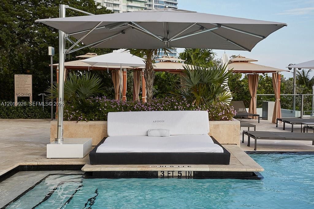 This image captures a luxurious poolside lounge area featuring a large, modern daybed positioned directly at the water's edge. A sleek, cantilevered umbrella provides shade over the white cushioned seating, which is branded with the name 'APOGEE'. In the background, lush tropical landscaping and additional cabanas create a resort-like atmosphere, emphasizing the high-end amenities of the property.